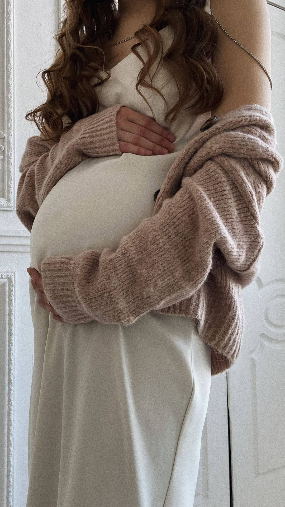 Woman 桌布 壁紙 — 一位孕婦穿著柔軟的米色連衣裙和厚實的針織開衫，輕輕地抱著她的肚子，靠在一扇白色鑲板門上。