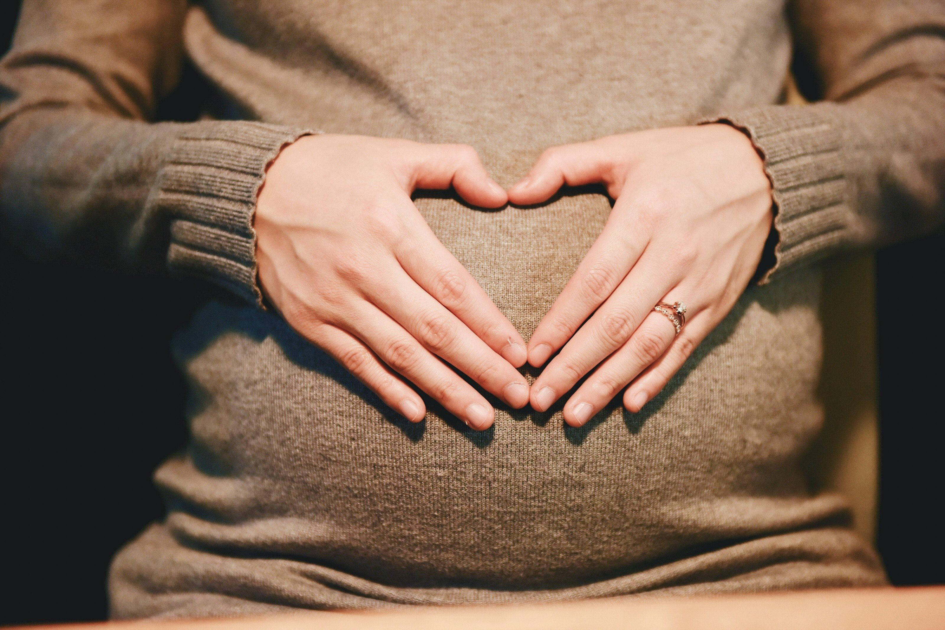 Pregnant 桌布 壁紙 — 一位孕婦穿著舒適的棕色毛衣，雙手在肚子上輕輕形成心形。