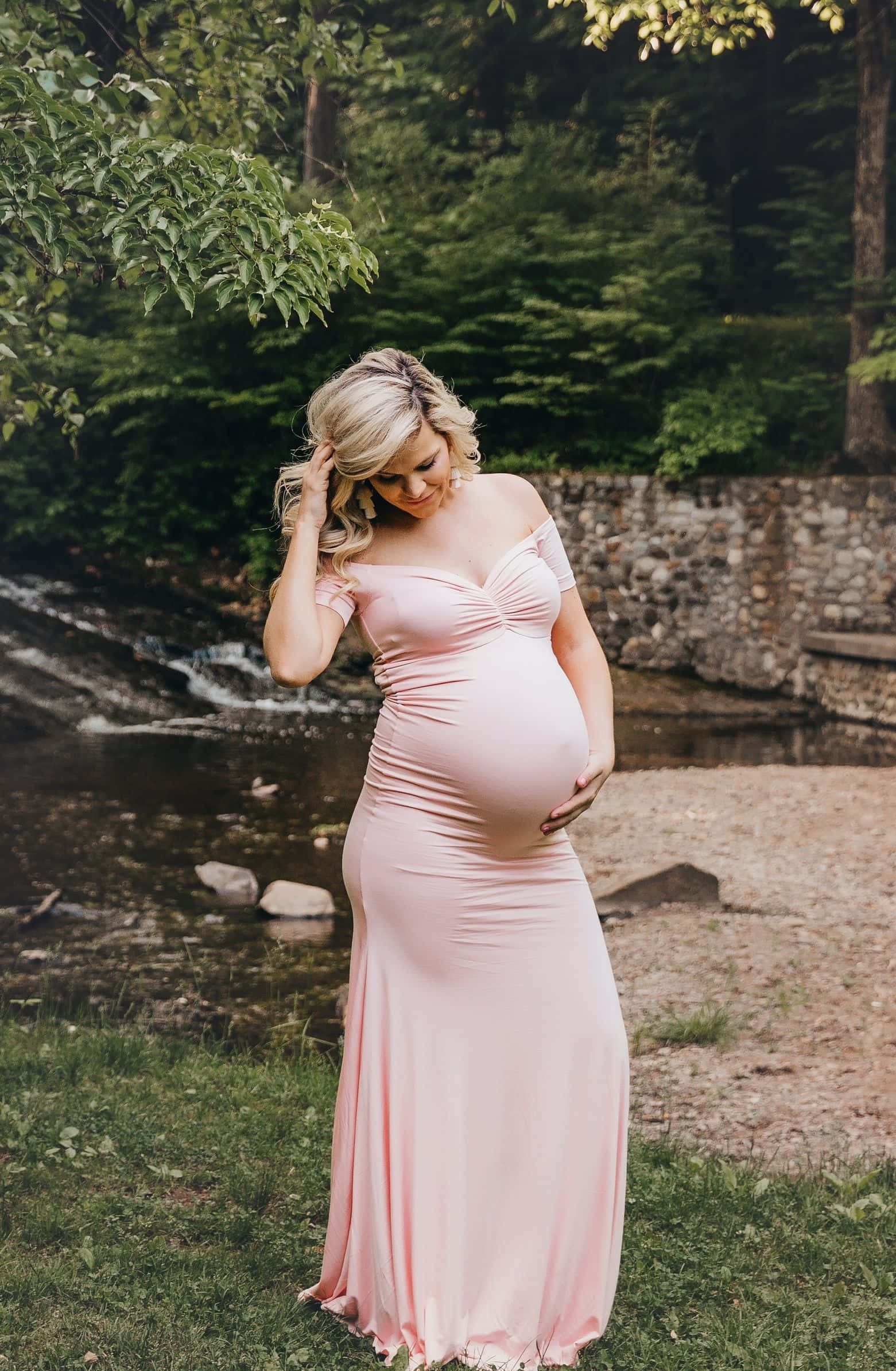 Pregnant 桌布 壁紙 — 一位孕婦穿著飄逸的粉紅色長袍，站在森林溪流旁，在斑駁的陽光下輕輕撫摸著肚子。