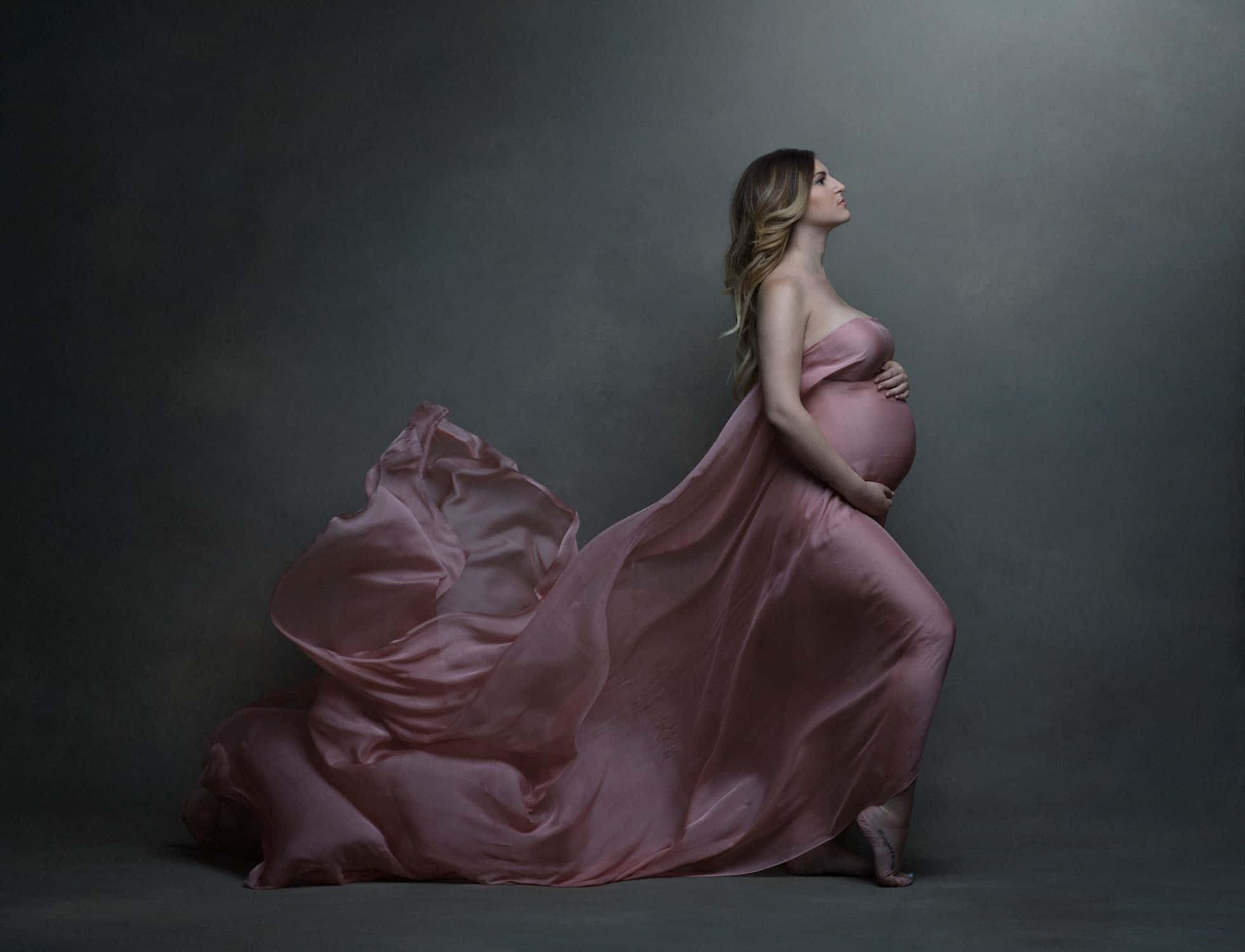 Pregnant 桌布 壁紙 — 一位孕婦身穿飄逸的粉紅色長袍，在深灰色背景下優雅地站立，雙手輕捧腹部，布料在她周圍飄揚。