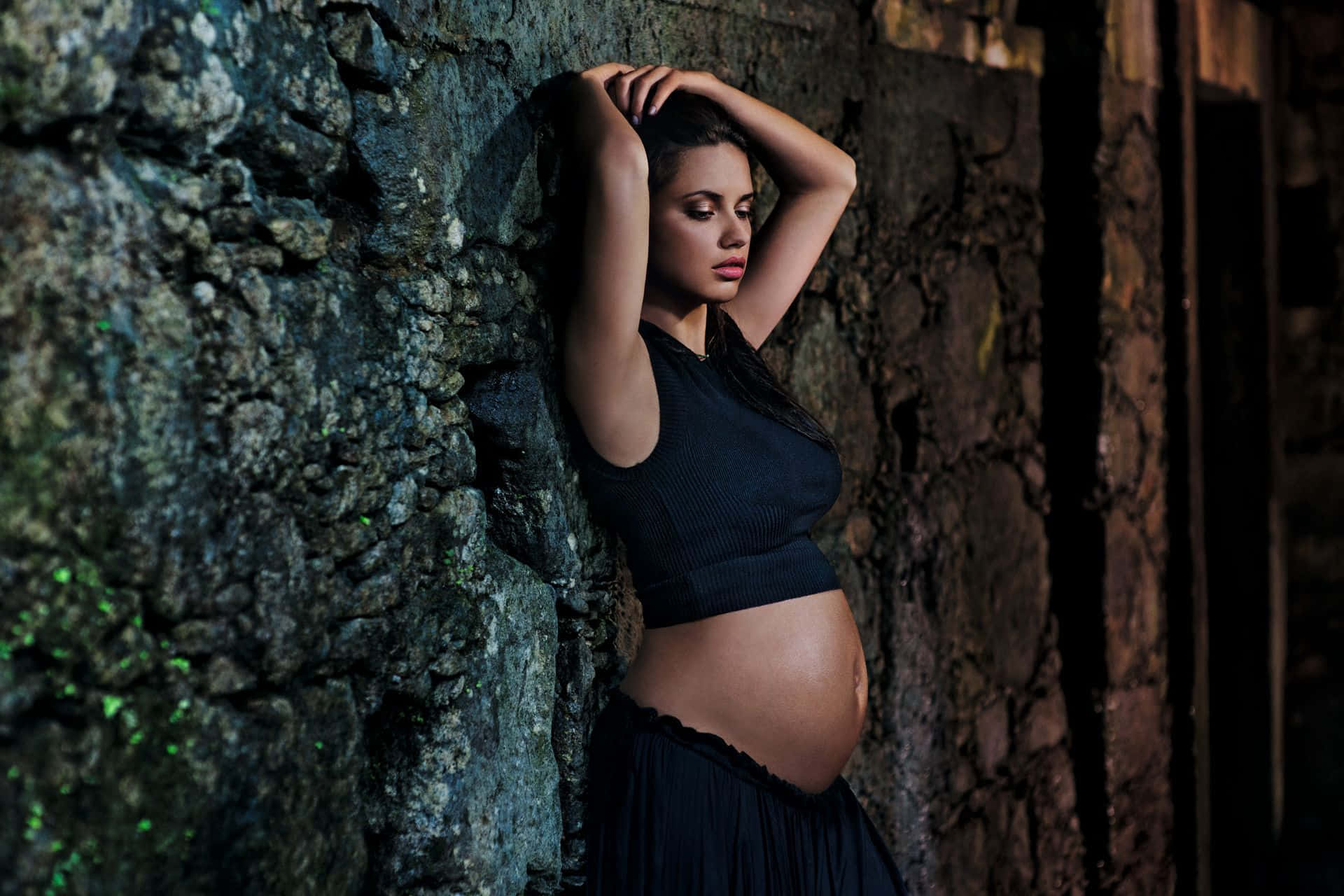 Pregnant 桌布 壁紙 — 一位孕婦身穿黑色露臍上衣，在昏暗簡樸的環境中靠著長滿青苔的石牆。