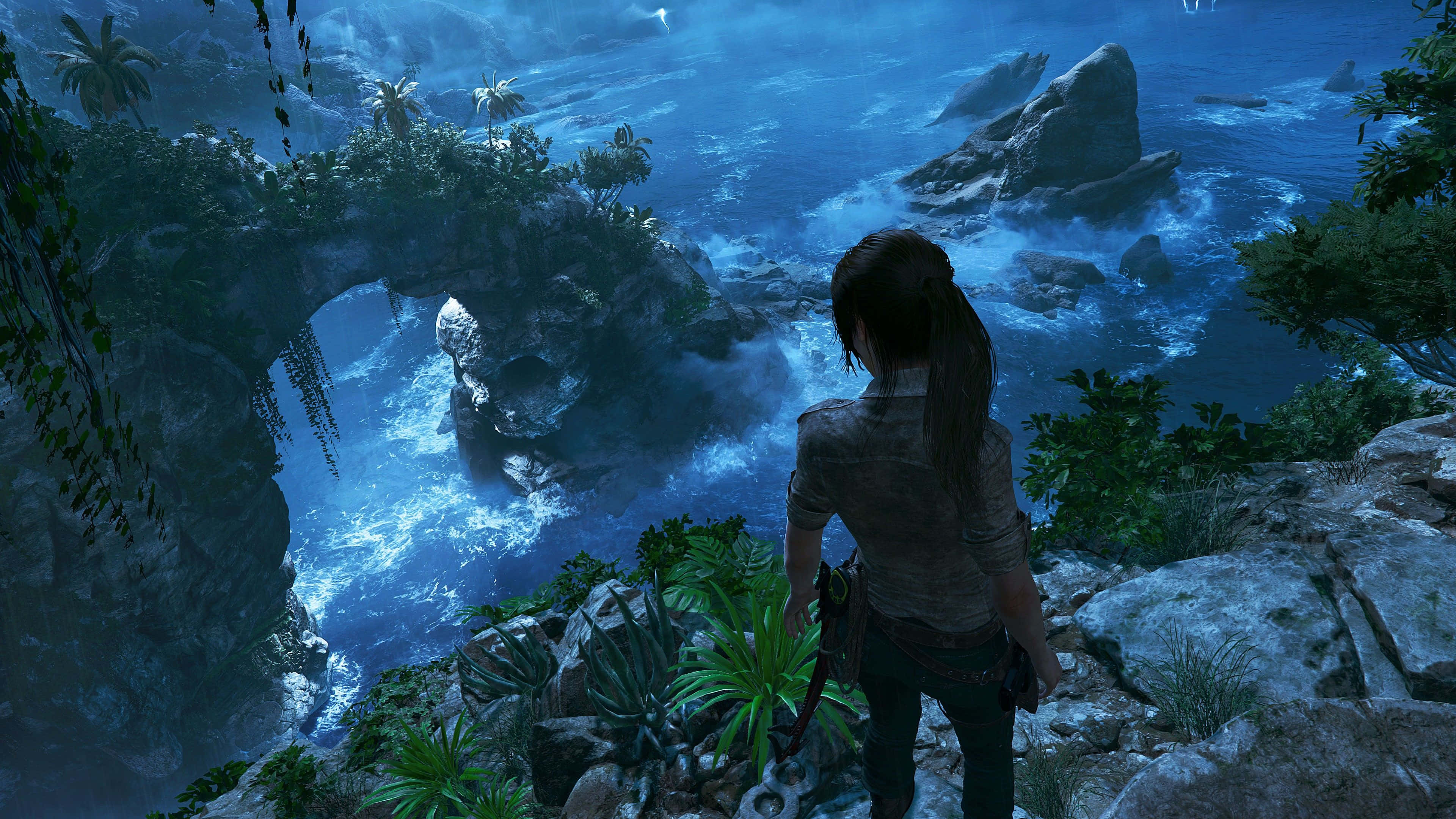 Lara Croft 桌布 壁紙 — 一位孤獨的冒險家站在岩石懸崖上，俯瞰著閃閃發光的藍色海洋，擁有天然石拱門和茂密的熱帶植被。