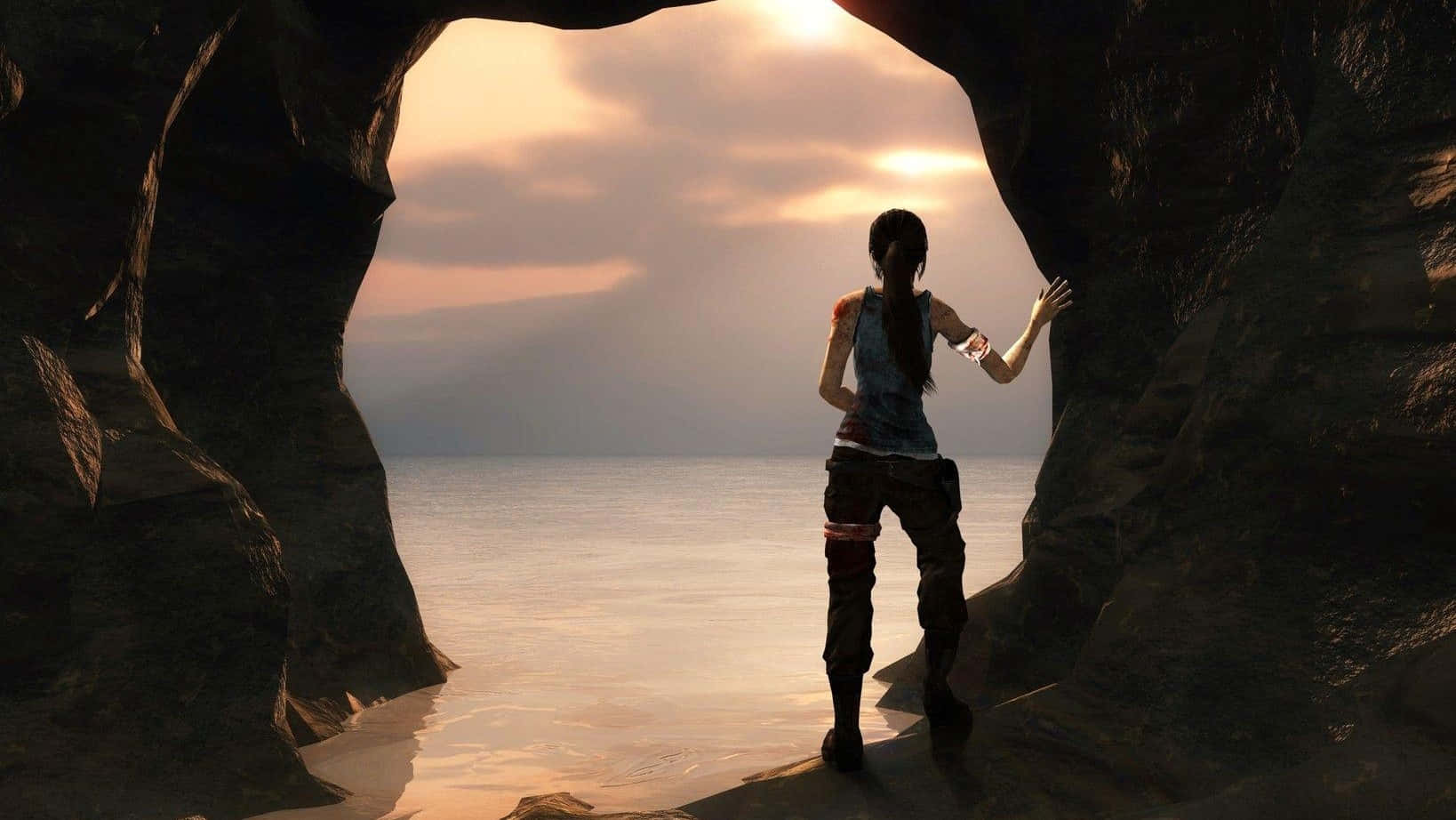 1080p Shadow Of The Tomb Raider 桌布 壁紙 — 一位孤獨的冒險家站在岩石洞穴入口，凝視著在燦爛日落天空下的寧靜海洋。