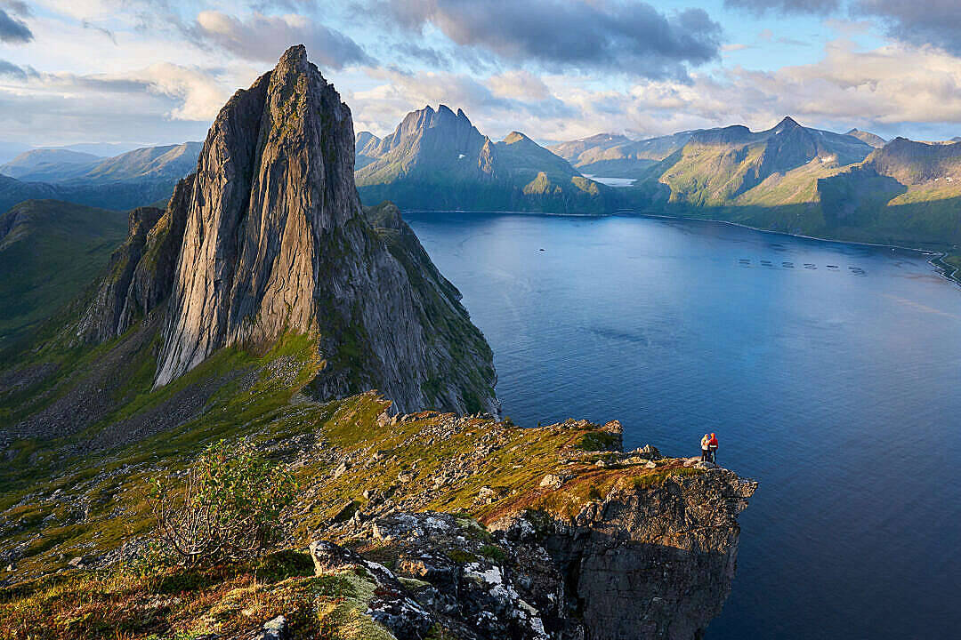 1920x1080 高畫質自然 桌布 壁紙 — 一位孤獨的徒步旅行者站在懸崖邊緣，俯瞰著廣闊的藍色峽灣，周圍環繞著高聳的綠色山脈，天空部分多雲。