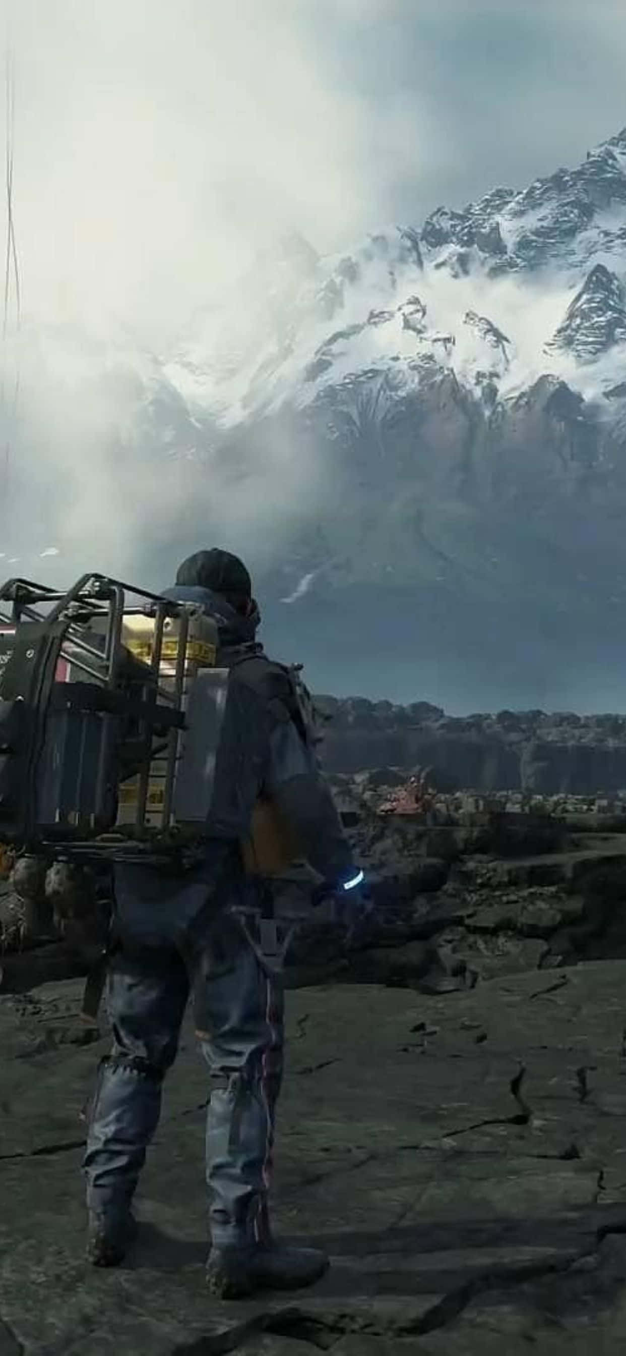 iPhone Xs Max Death Stranding 桌布 壁紙 — 一位孤獨的探險家穿著堅固裝備，站在岩石露頭上，凝視著陰天天空下高聳的雪山山脈。