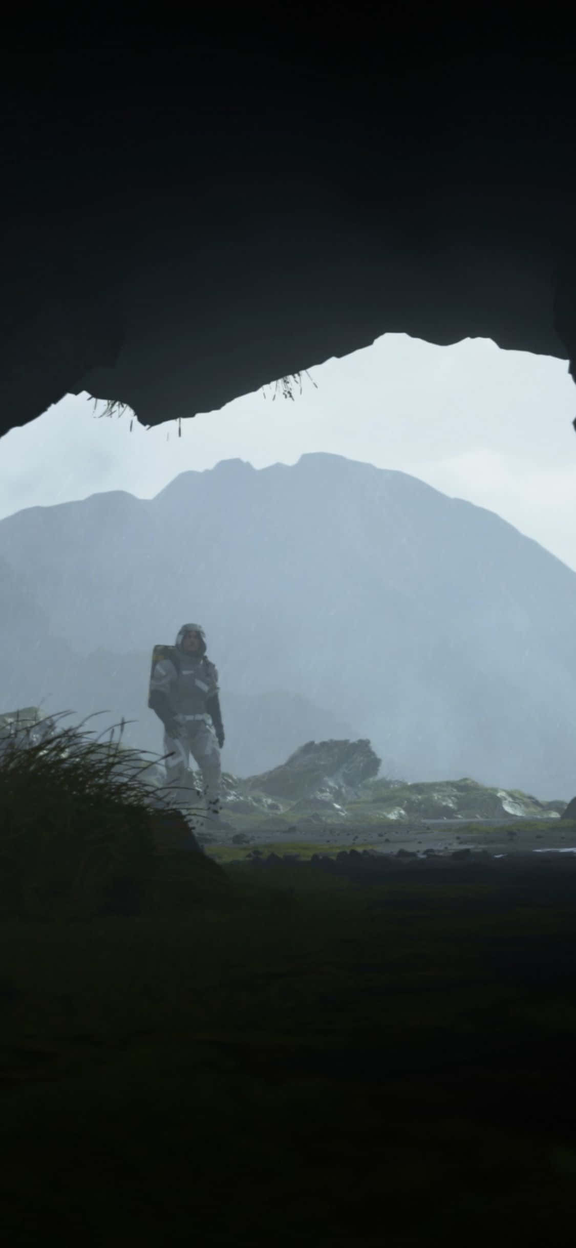 iPhone Xs Death Stranding 桌布 壁紙 — 一位孤獨的探險家穿著未來主義套裝站在洞穴邊緣，凝視著陰天下的霧氣山脈。