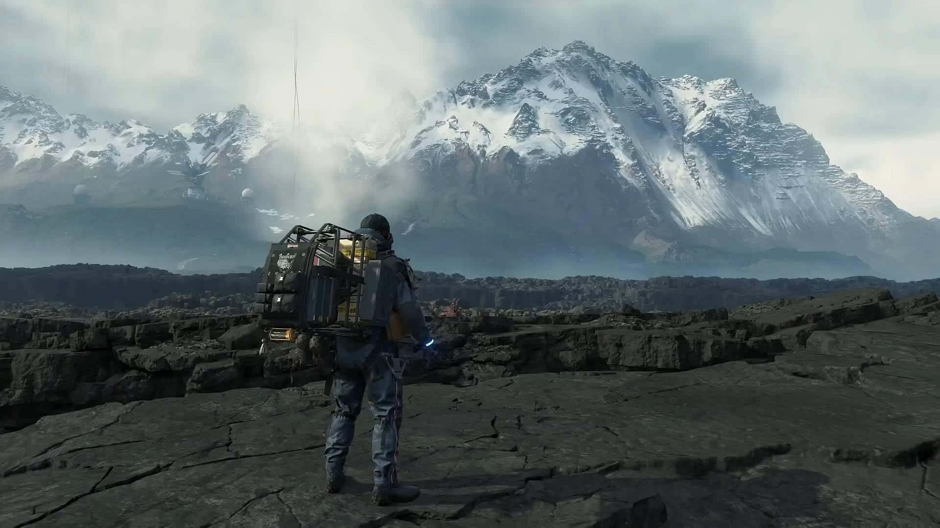 Death Stranding Pc 桌布 壁紙 — 一位孤獨的探險家穿著未來裝備，站在岩石平原上，凝視著陰雲天空下高聳的雪山。
