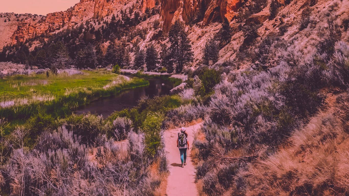 Traveler 桌布 壁紙 — 一位孤獨的揹著背包的徒步旅行者沿著一條蜿蜒的泥土小徑，在一條平靜的河流旁，穿過崎嶇的峽谷景觀。