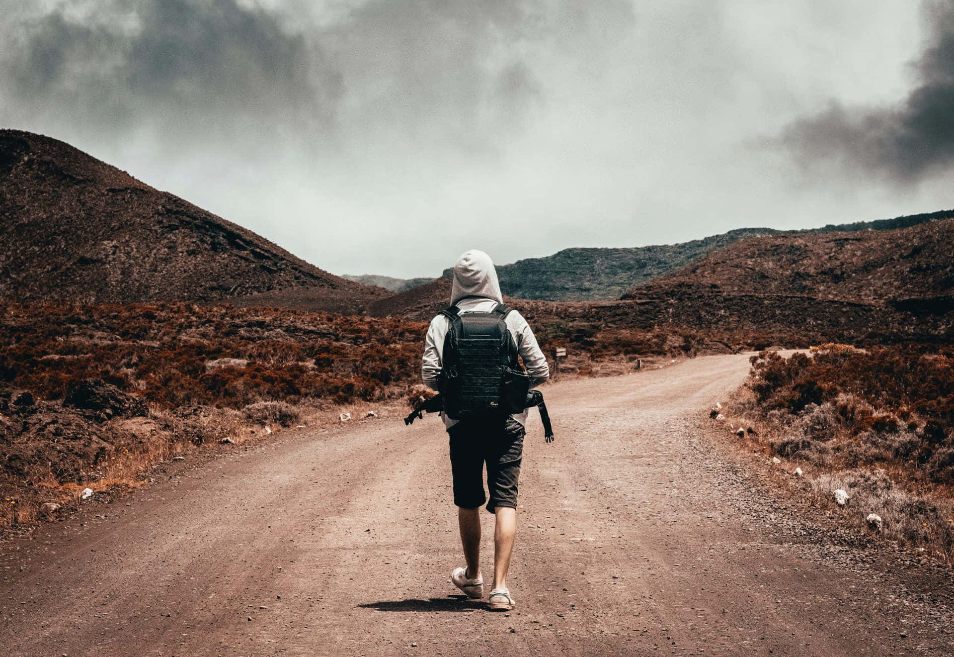 Hiking 桌布 壁紙 — 一位孤獨的揹著背包的遠足者，在陰天之下，沿著一條蜿蜒的泥路穿過一片貧瘠、岩石遍佈的景觀。