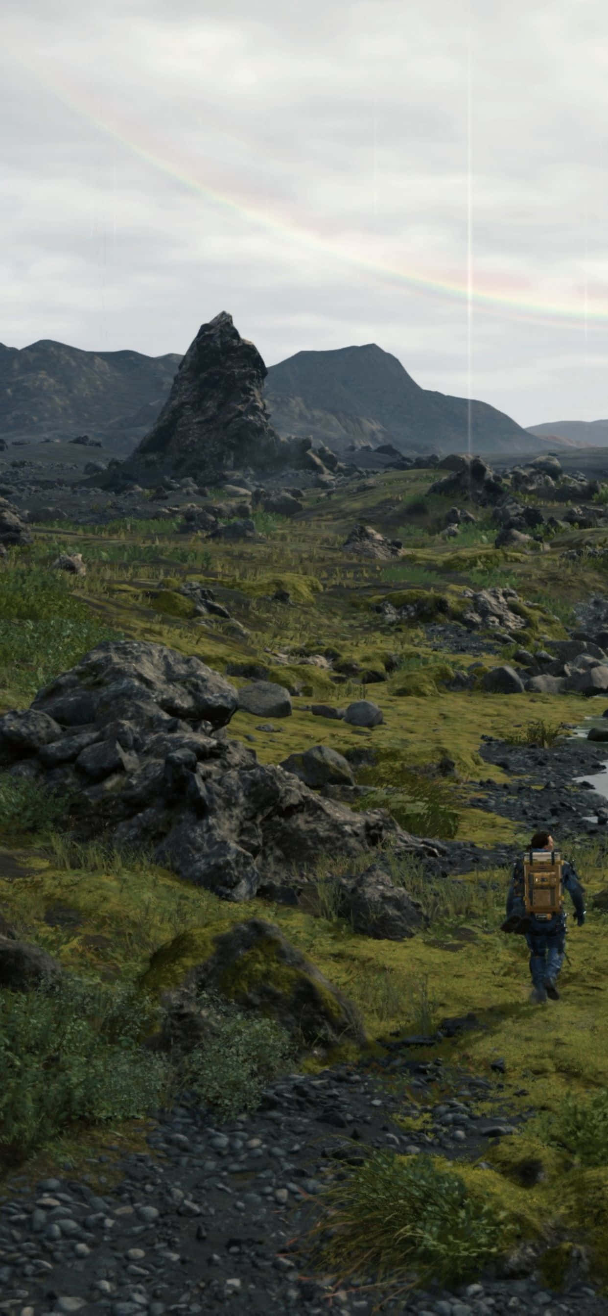 iPhone Xs Max Death Stranding 桌布 壁紙 — 一位孤獨的揹著背包的遠足者，在陰天之下走過長滿苔蘚的岩石景觀，頭頂有一道淡淡的彩虹拱起。