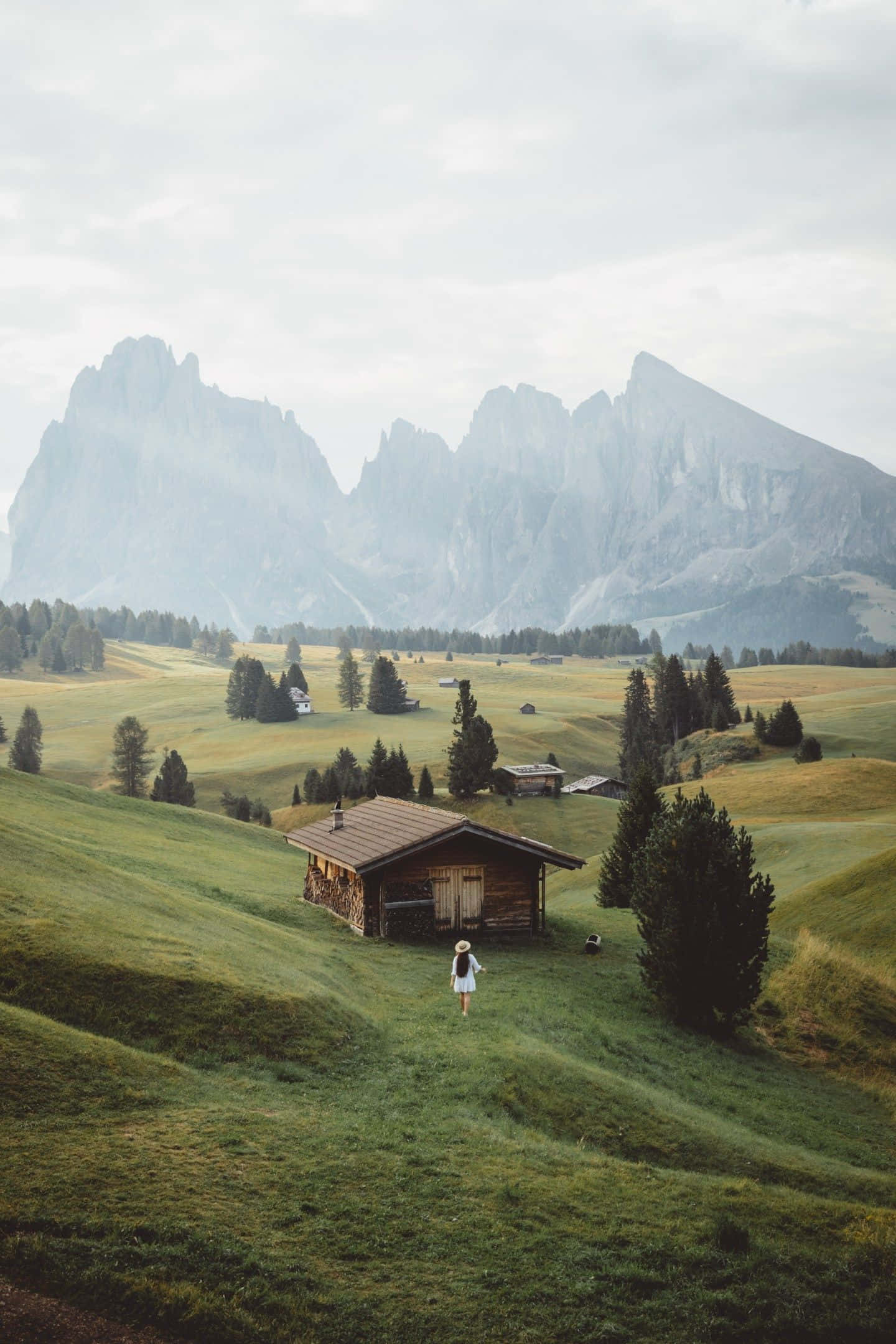 Beautiful Nature 桌布 壁紙 — 一位孤獨的旅人走向一座坐落在廣闊綠色山谷中的質樸木屋，遠處霧氣繚繞，高聳崎嶇的山巒若隱若現。