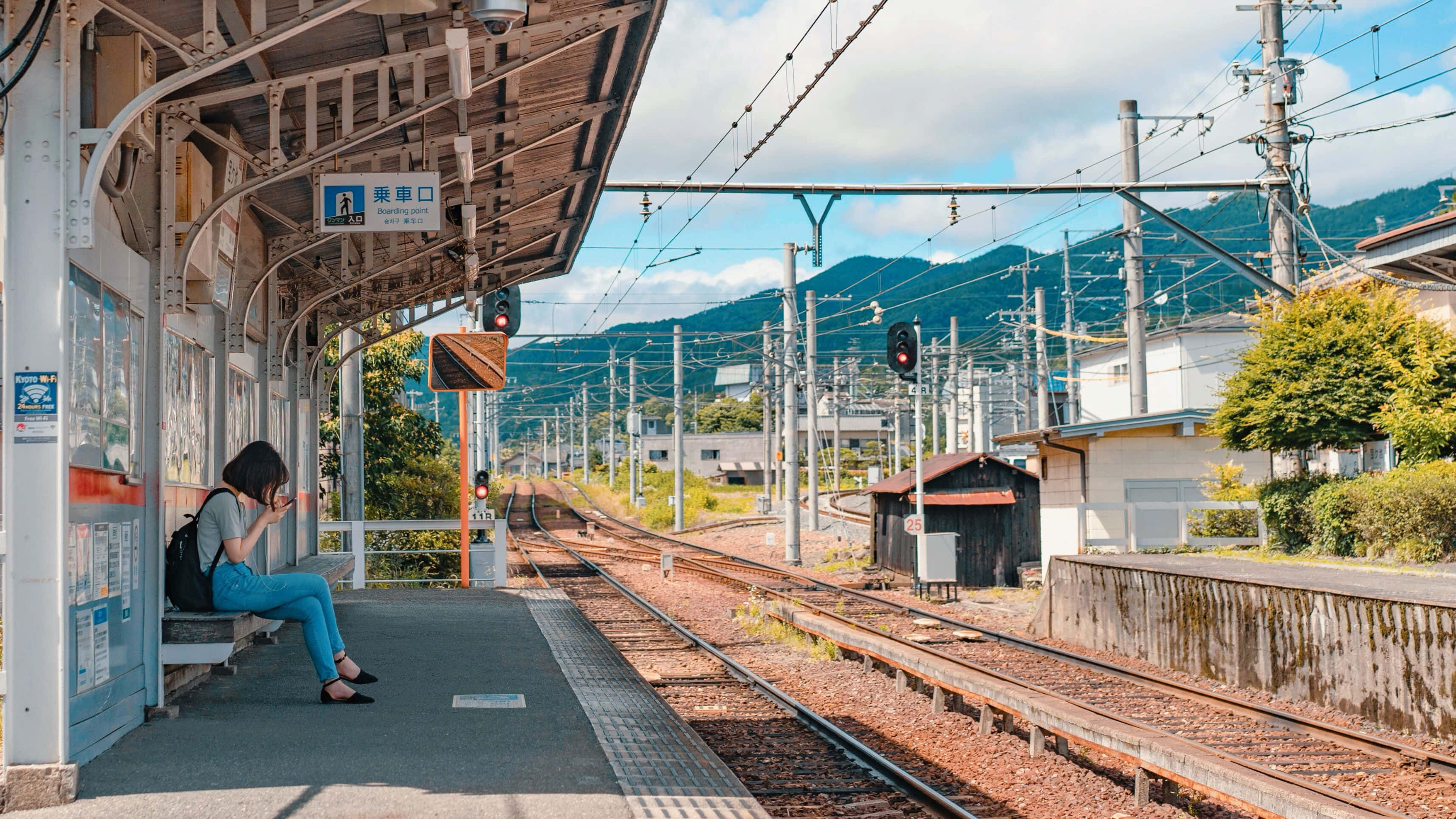 Train Station 桌布 壁紙 — 一位孤獨的旅客坐在火車站的木製簷篷下的長椅上，鐵軌在明亮的藍天下延伸向遠處的綠色山丘。