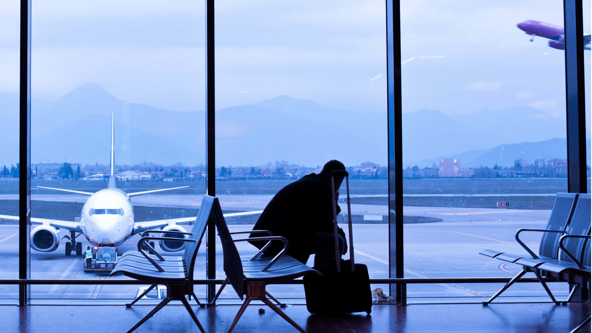 Flight 桌布 壁紙 — 一位孤獨的旅客帶著行李箱坐在機場航站樓，凝視著停泊的飛機和遠山，背景是清冷的藍色暮色天空。
