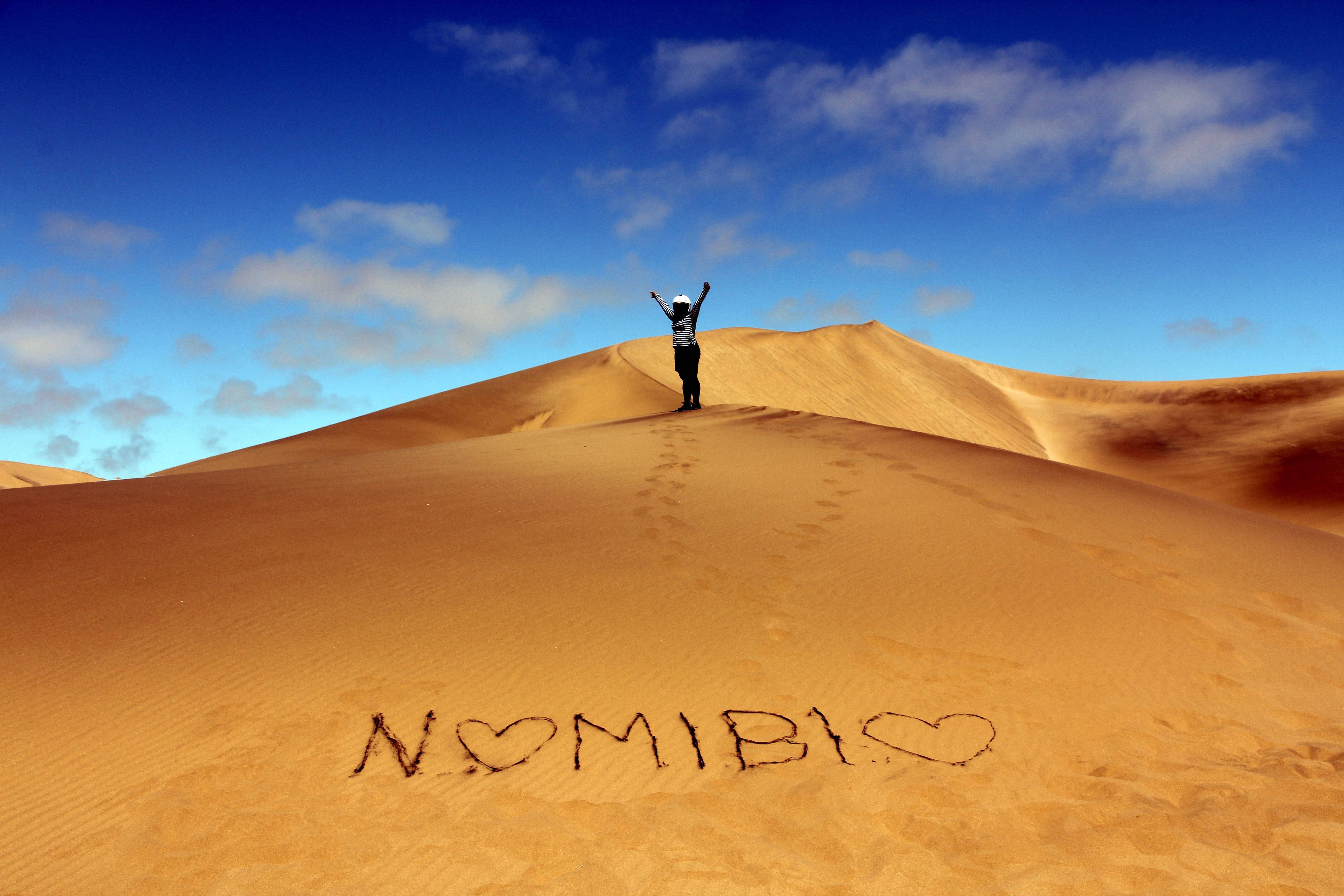 Namibia 桌布 壁紙 — 一位孤獨的旅行者勝利地站在金色沙丘頂端，雙臂高舉，頭頂是廣闊的藍天，點綴著蓬鬆的白雲。