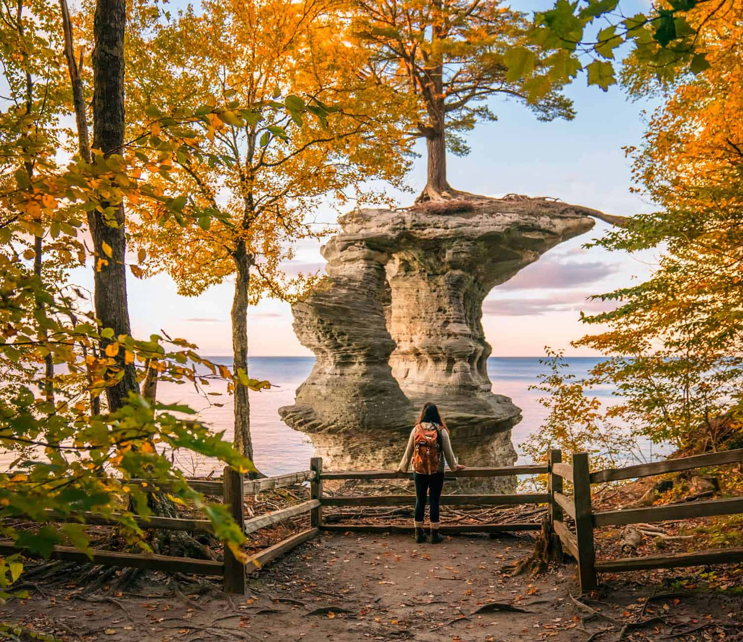 Michigan 桌布 壁紙 — 一位孤獨的旅行者站在一座頂端有樹木的 towering 岩石 formation 前，金色的秋葉框住了寧靜的湖邊景色。