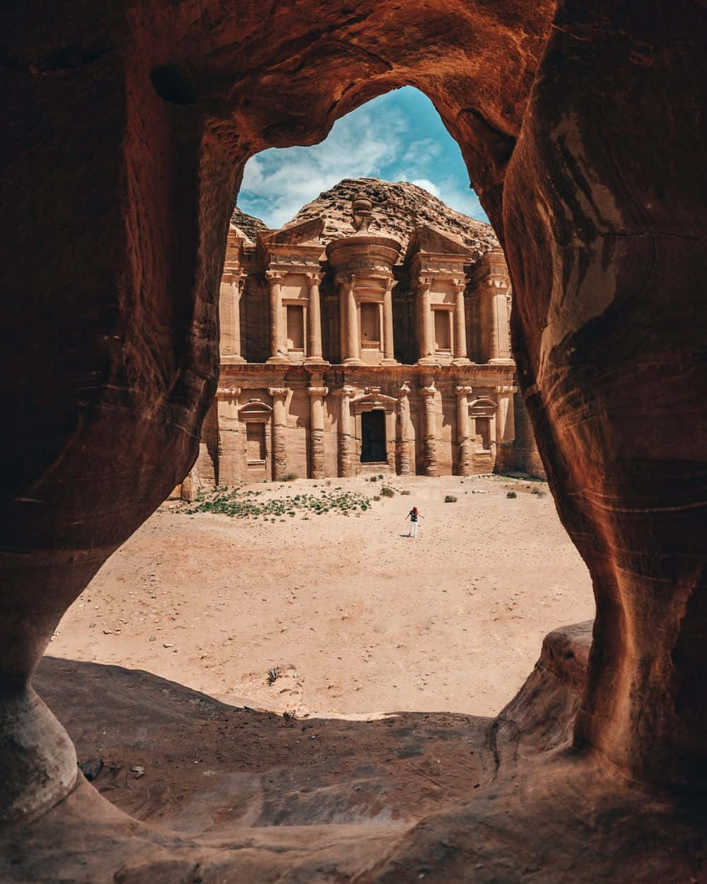 Jordan 桌布 壁紙 — 一位孤獨的旅行者站在古老的玫瑰紅色佩特拉寶庫前，由天然岩石拱門框住，背景是明亮的藍天。