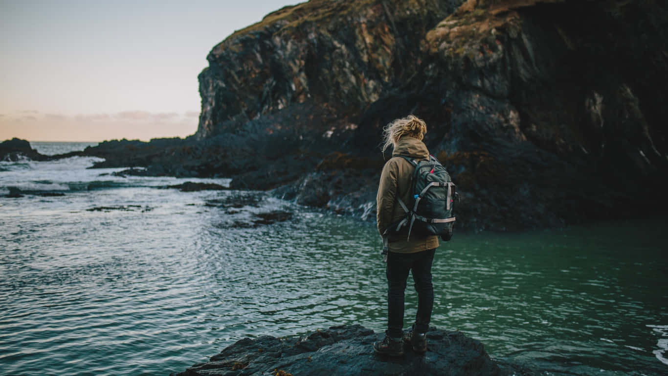 Traveler 桌布 壁紙 — 一位孤獨的旅行者背著背包站在岩石峭壁上，俯瞰黃昏時平靜的海面。