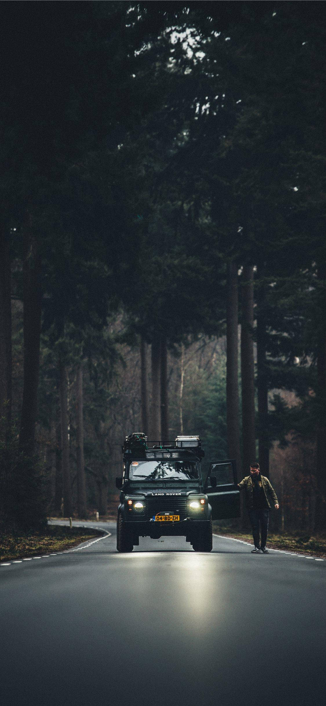X 桌布 壁紙 — 一位孤獨的旅行者走在車頭燈照亮的黑暗森林路上，旁邊停著一輛綠色SUV。