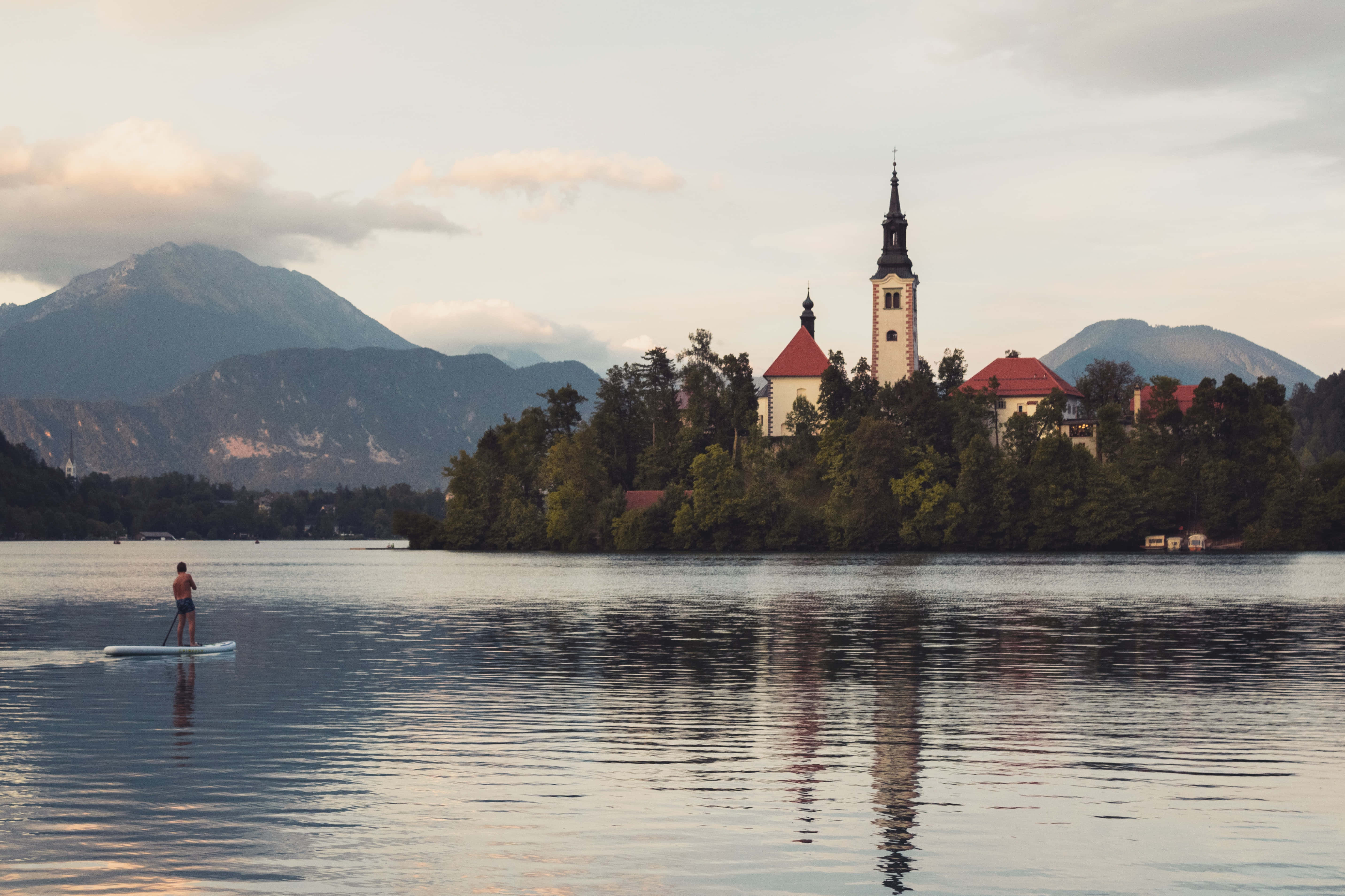Slovenia 桌布 壁紙 — 一位孤獨的槳板划手在平靜的湖面上滑行，前往一座島嶼教堂，教堂有高聳的尖塔，背景是遠山，正值黃金時刻。