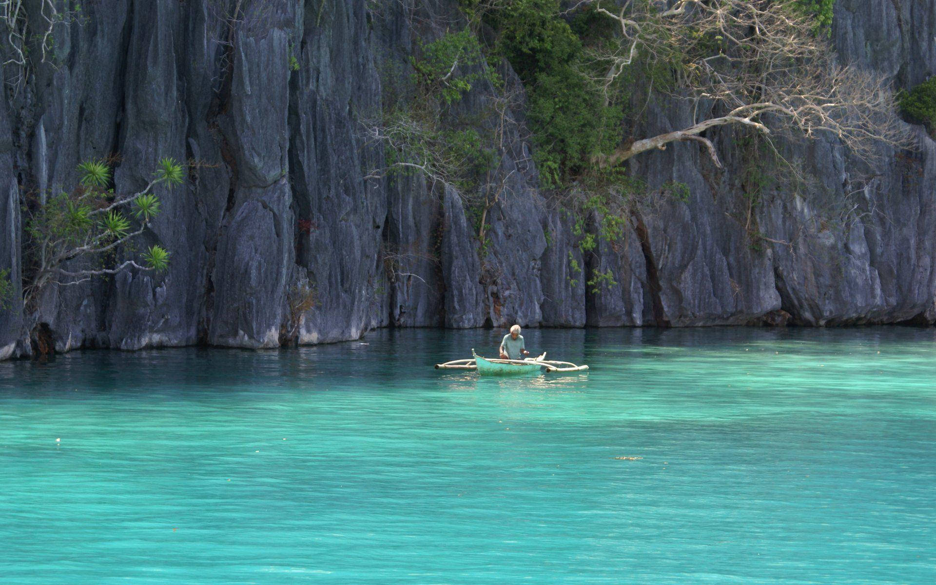 Philippines 桌布 壁紙 — 一位孤獨的漁夫划著小船，穿過晶瑩剔透的碧綠海水，旁邊是高聳的深色石灰岩懸崖。