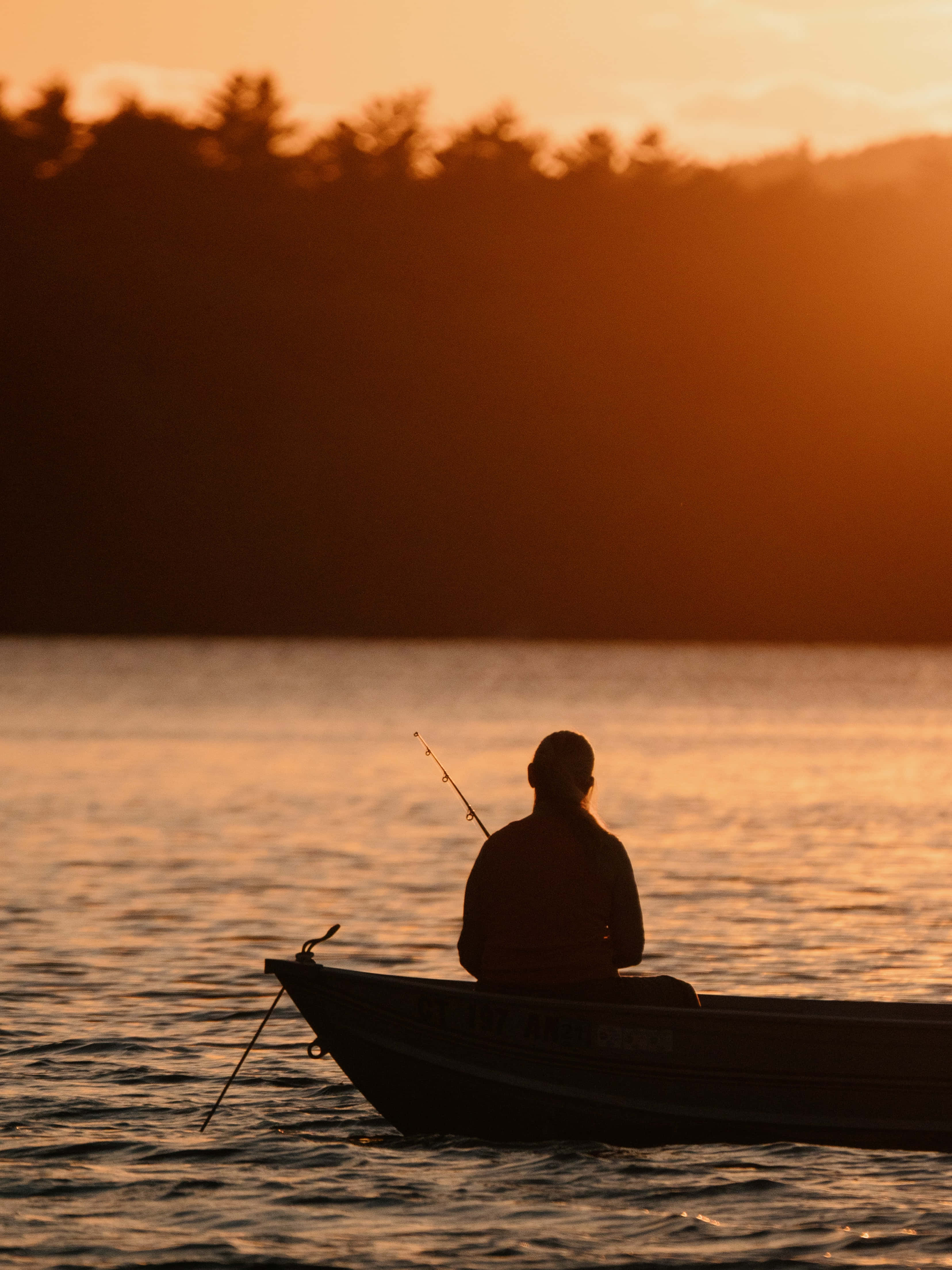 Fishing Phone 桌布 壁紙 — 一位孤獨的漁夫坐在小船中，平靜的水面上夕陽將溫暖的橙色光芒灑向遠處的樹線。