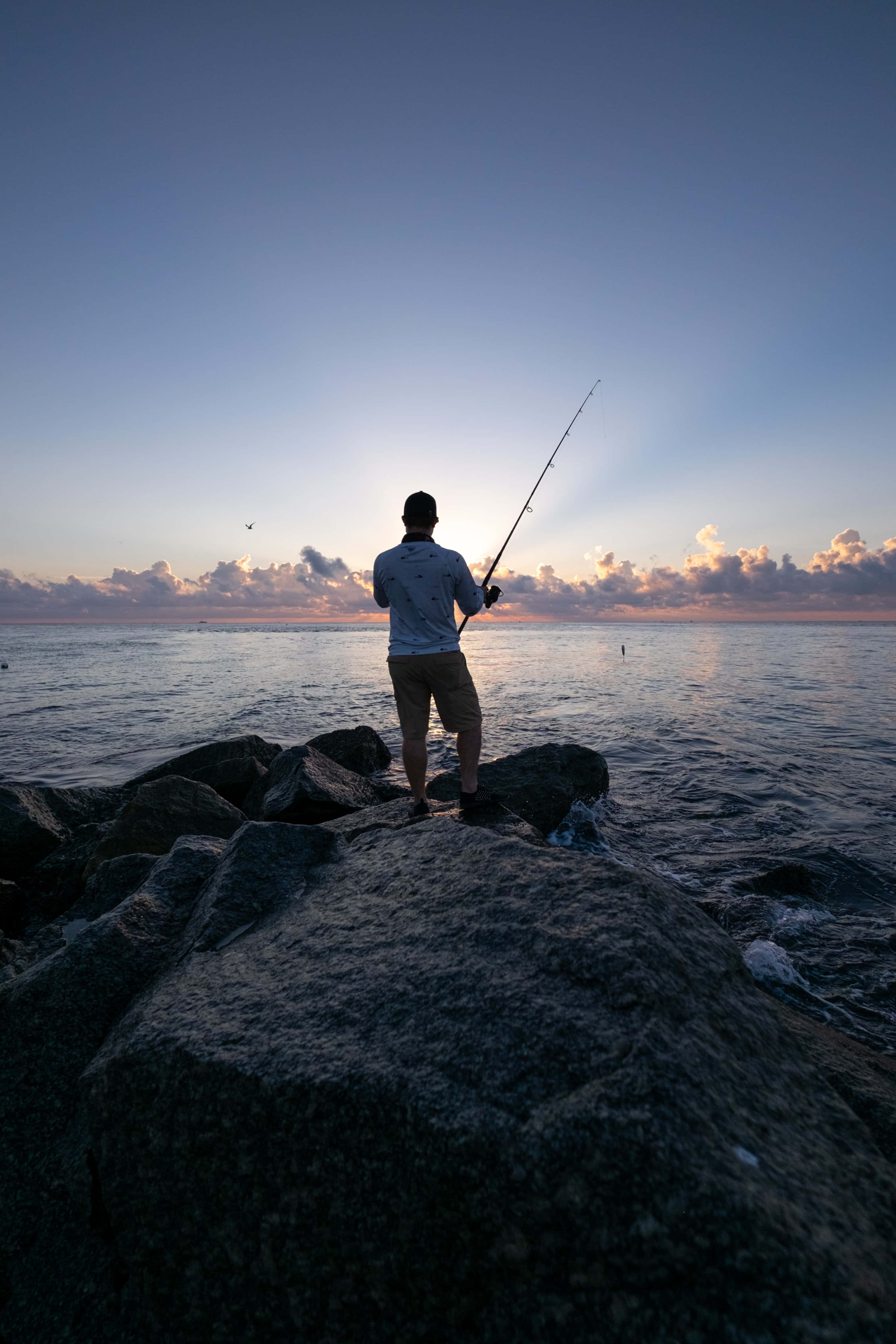 Fishing Phone 桌布 壁紙 — 一位孤獨的漁夫站在岩石海岸上，在金色日落時將釣線拋入平靜的大海。