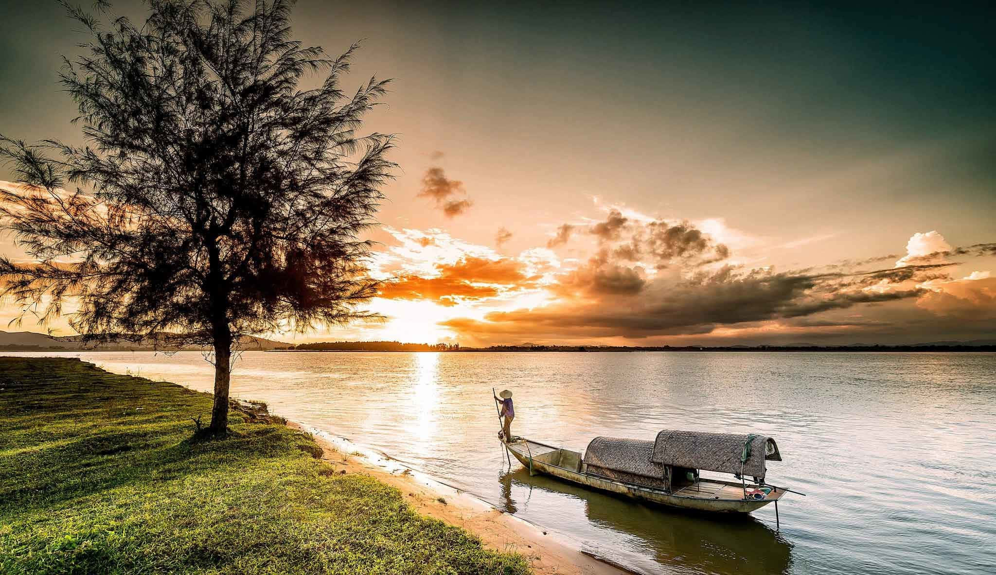 越南 桌布 壁紙 — 一位孤獨的漁夫站在木船上，夕陽西下，在火紅的天空和寧靜的水面上形成剪影，旁邊是長滿草的岸邊，有一棵孤樹。