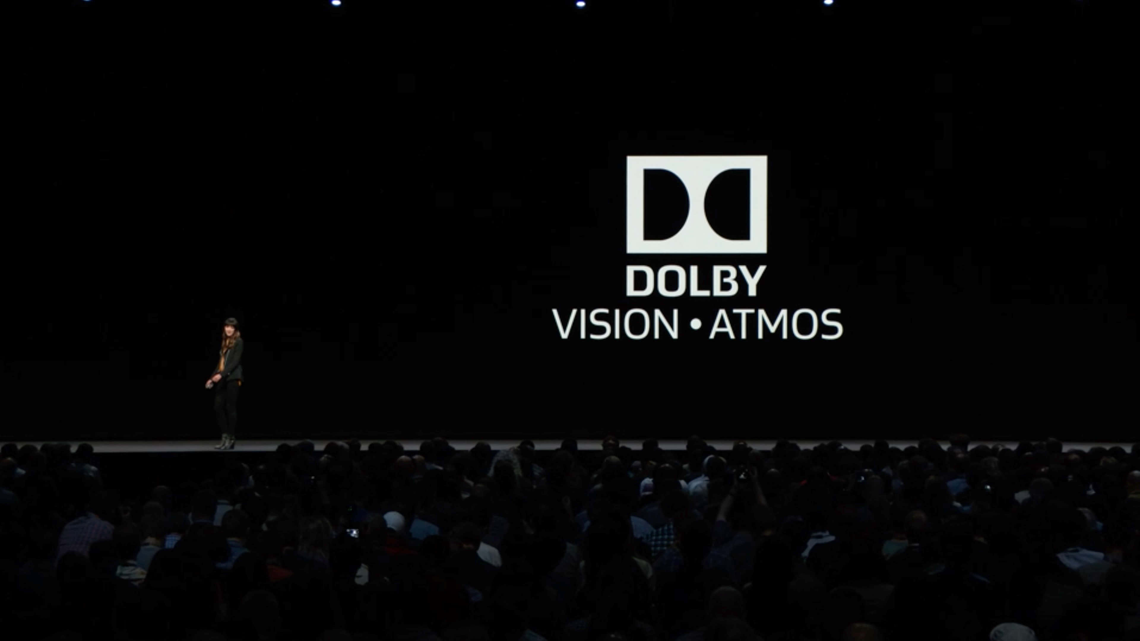 4K Dolby Vision 桌布 壁紙 — 一位孤獨的演講者站在黑暗的舞台上，面對著一個巨大的螢幕，螢幕上顯示著Dolby Vision Atmos標誌，面向一片剪影般的觀眾。