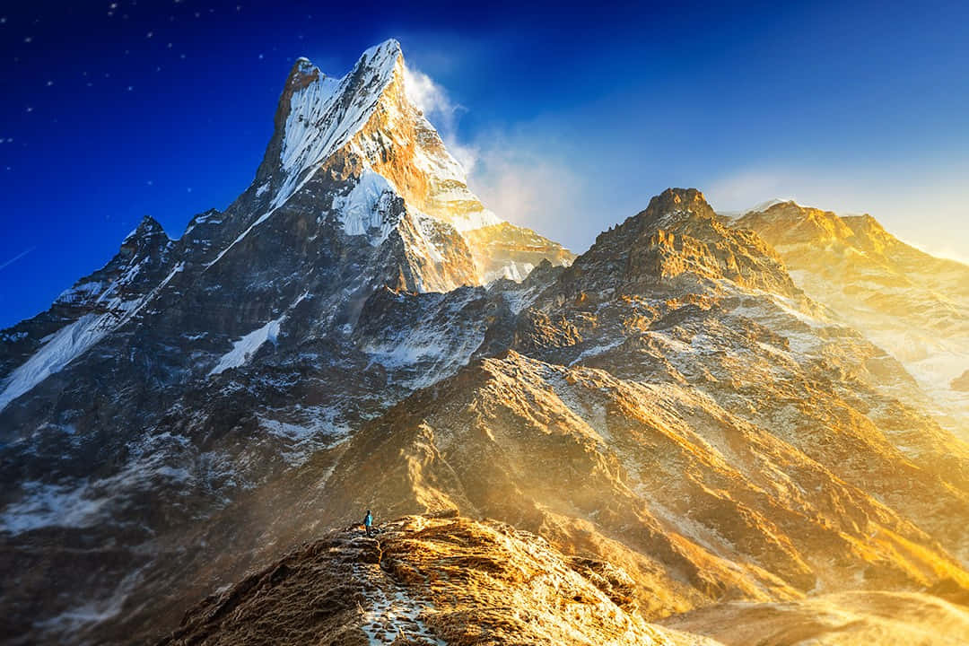 山景 桌布 壁紙 — 一位孤獨的登山者站在崎嶇的山脊上，金色的日出光芒沐浴著鋸齒狀的山峰，映襯著繁星點點的藍天。