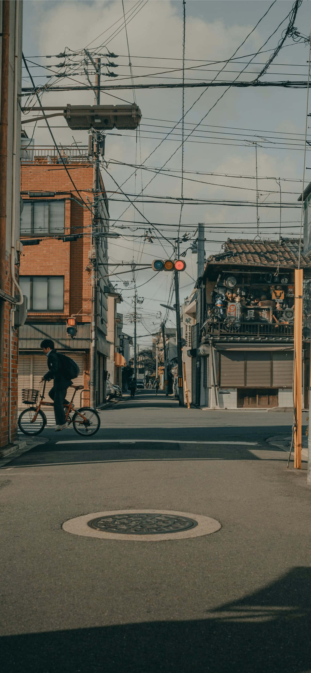 Japanese iPhone 桌布 壁紙 — 一位孤獨的自行車騎士在朦朧的天空下，沿著一條兩旁是磚砌建築和纏繞電線的狹窄日本街道騎行。
