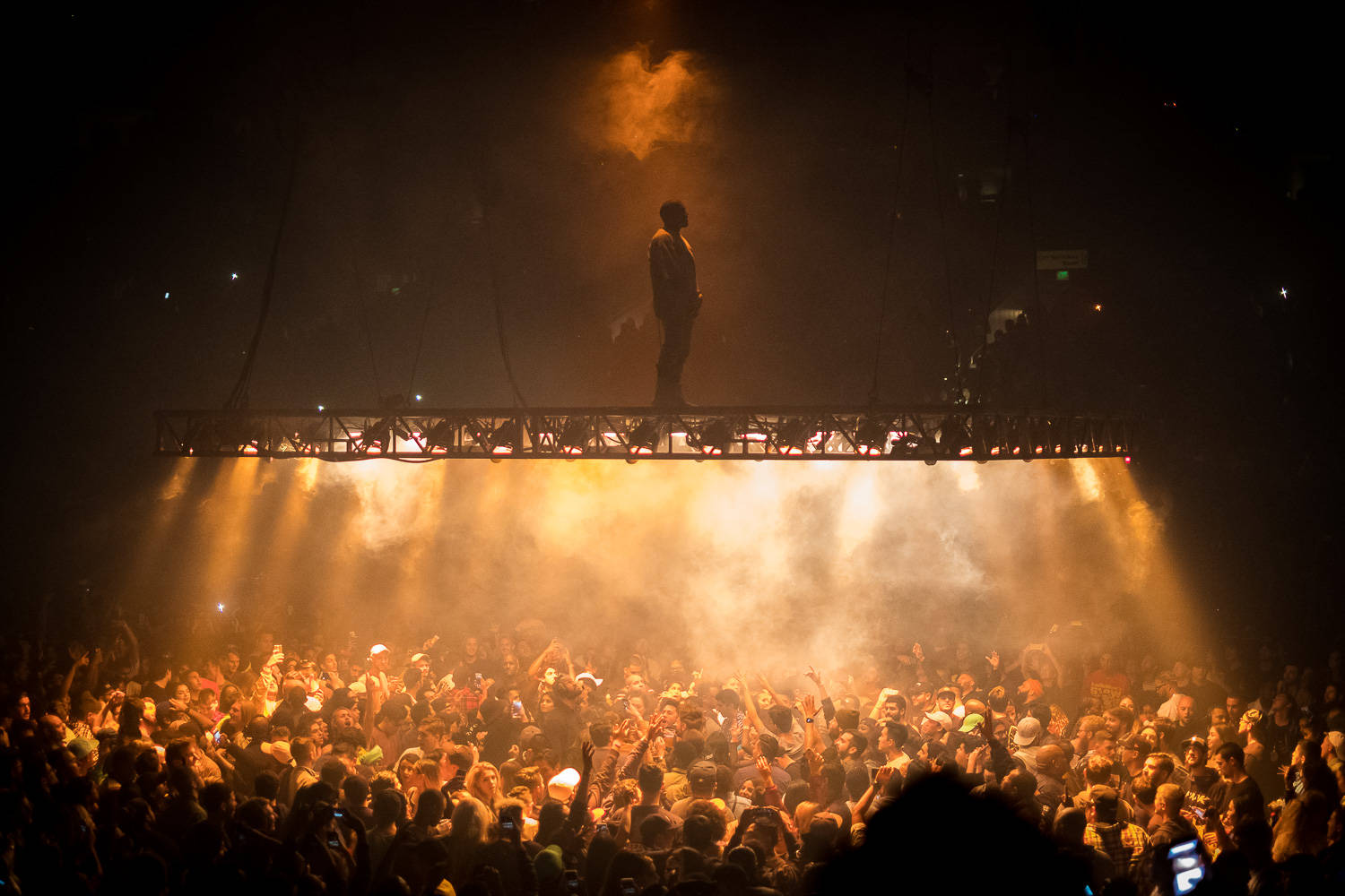 Kanye 桌布 壁紙 — 一位孤獨的表演者以剪影站在懸掛的舞台上，下方是沐浴在金色舞台燈光中的喧鬧人群。