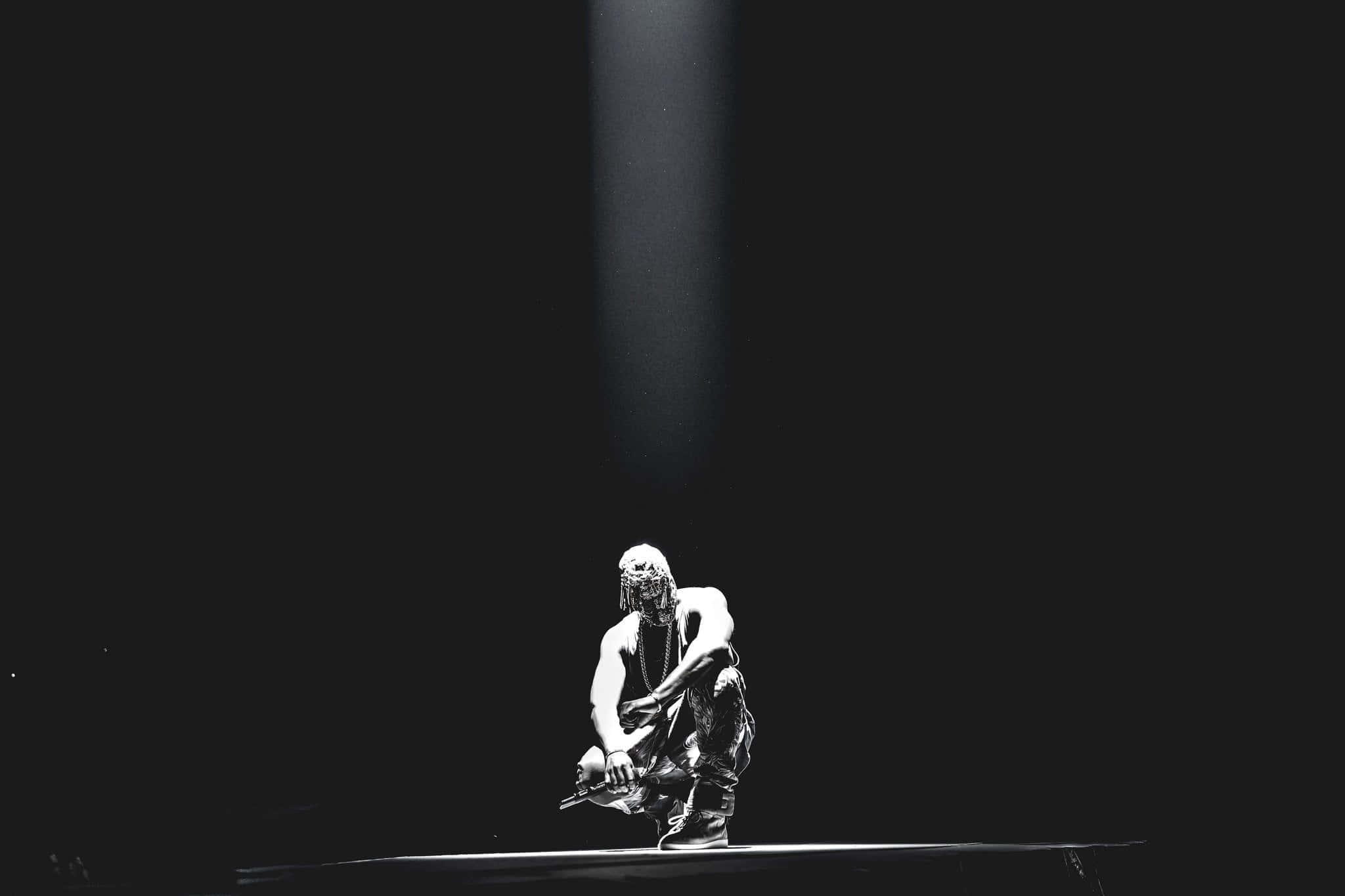 Kanye West Yeezus 桌布 壁紙 — 一位孤獨的表演者在黑暗舞台上的單一聚光燈下蹲伏，營造出戲劇性和強烈的氛圍。