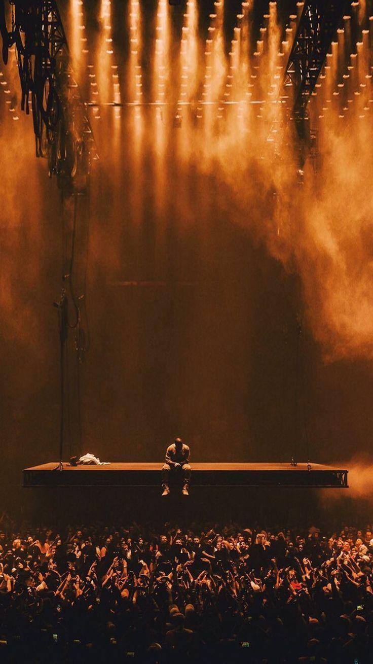 Kanye 桌布 壁紙 — 一位孤獨的表演者坐在懸浮舞台上，沐浴在金黃燈光中，下方是歡呼的粉絲海洋。