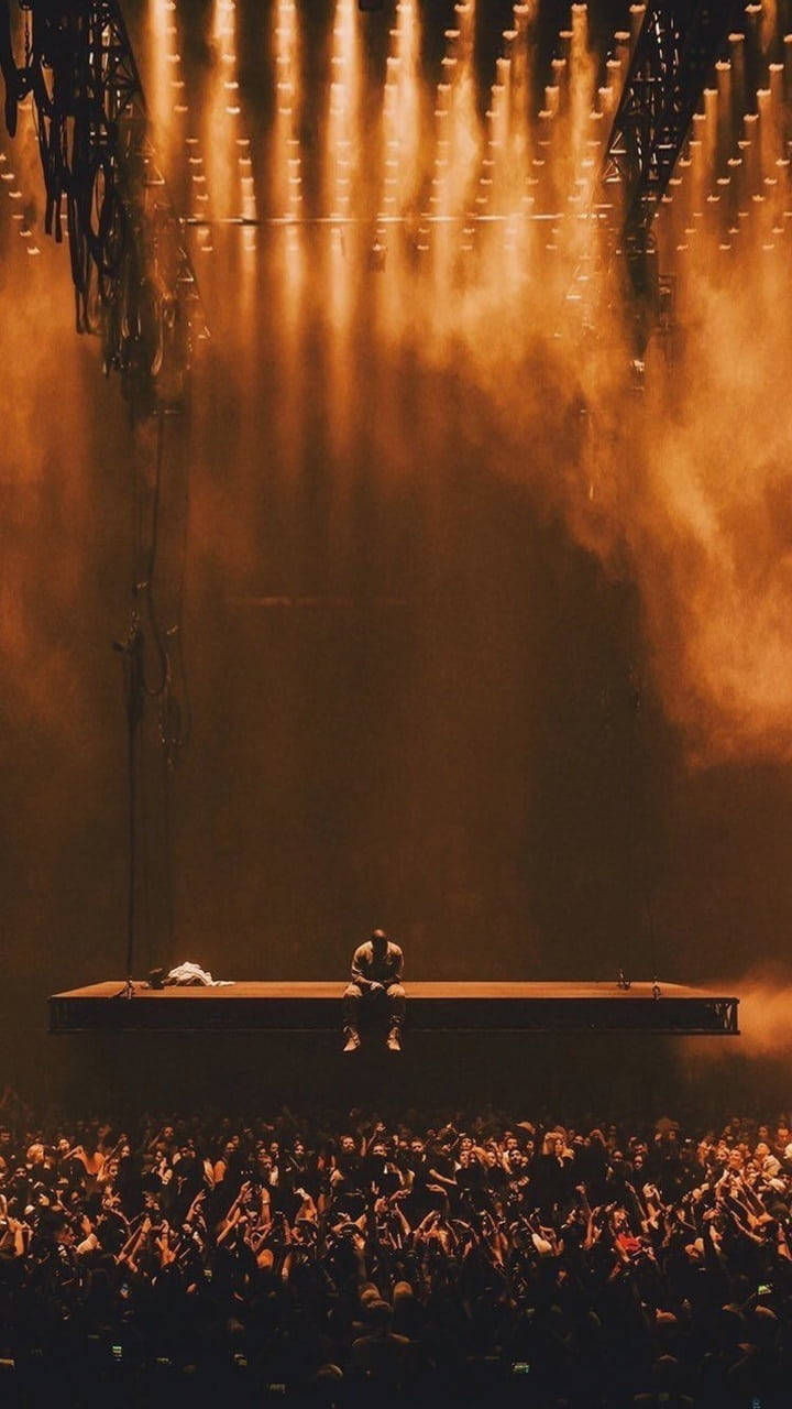 Kanye West Ye 桌布 壁紙 — 一位孤獨的表演者獨自坐在高台上，沐浴在金色聚光燈下，下方是歡呼的粉絲海洋。