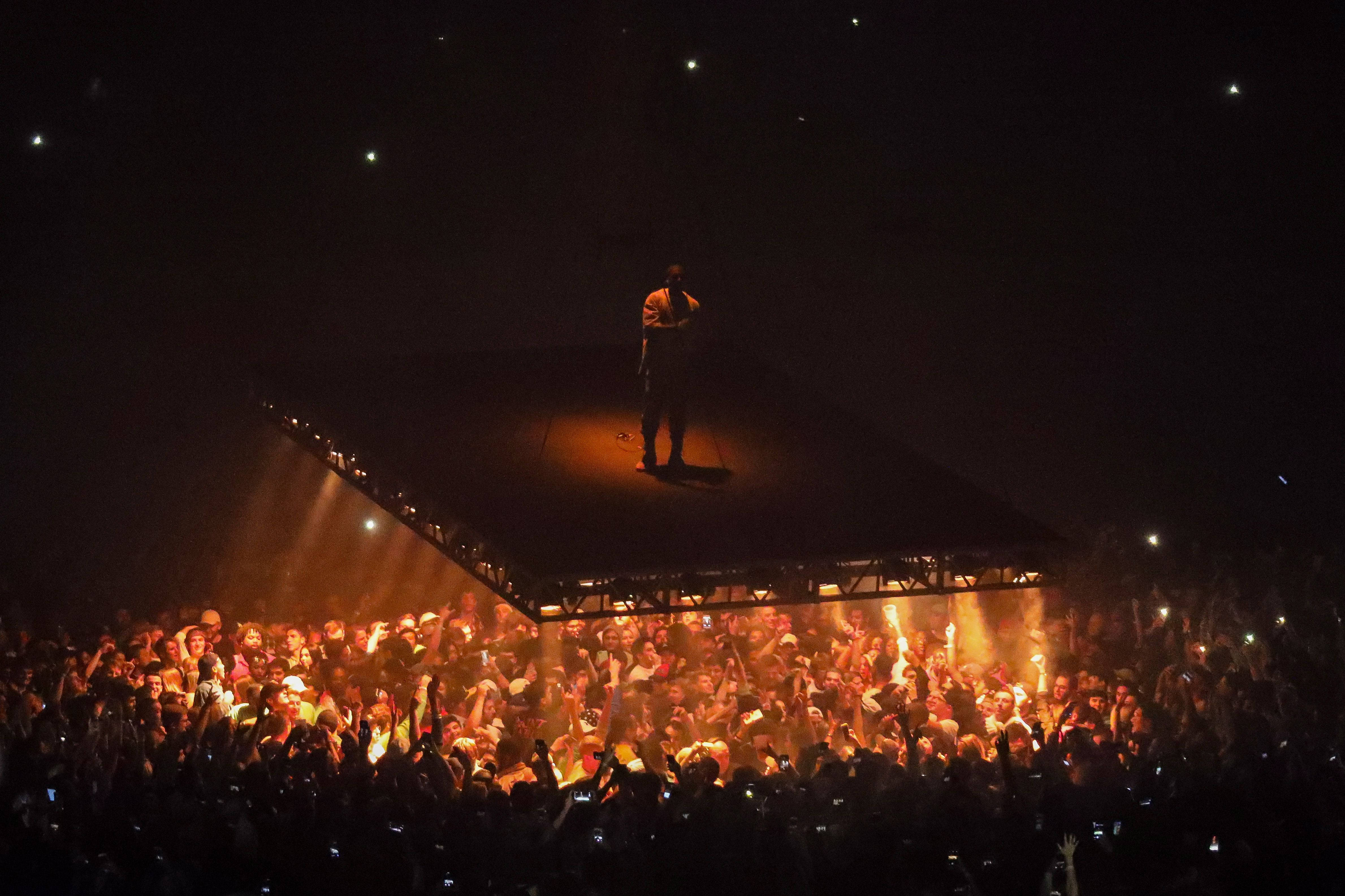 Kanye 桌布 壁紙 — 一位孤獨的表演者獨自站在黑暗的舞台上，沐浴在溫暖的聚光燈下，而下面的一片粉絲齊聲舉起發光的手機。