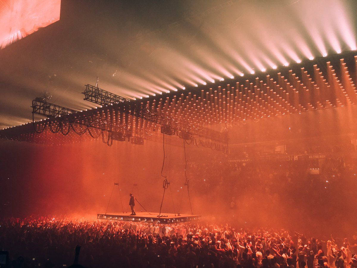 kanye west saint pablo 桌布 壁紙 — 一位孤獨的表演者站在一個漂浮的舞台上，沐浴在熾熱的橙色燈光中，下方是大型音樂會上咆哮的人群。