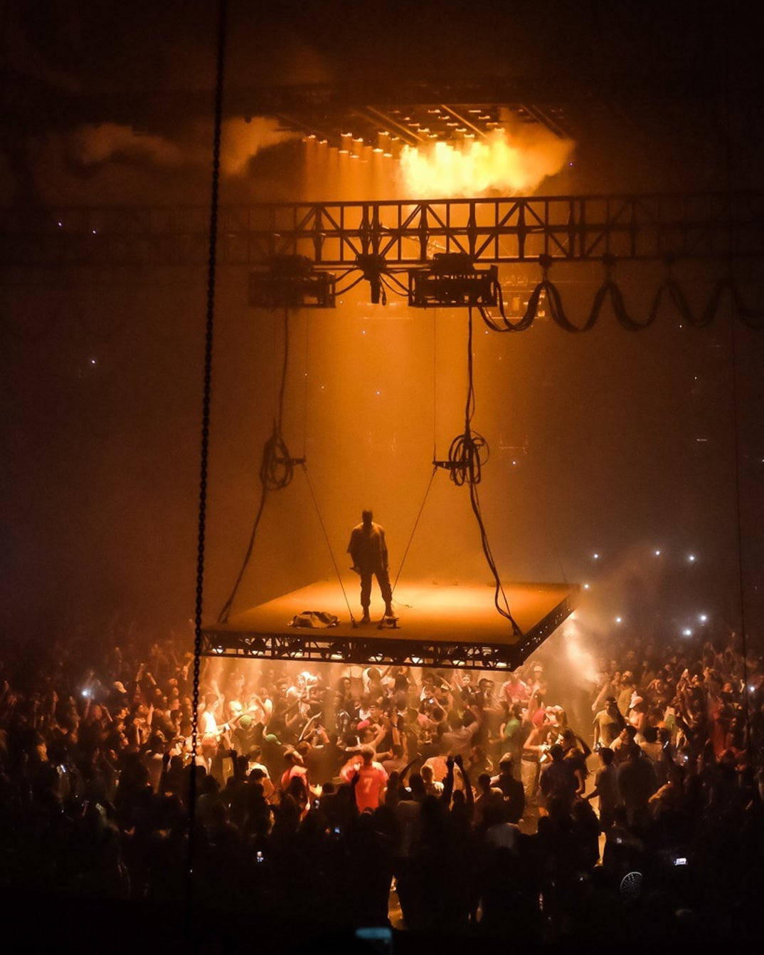 kanye west android 桌布 壁紙 — 一位孤獨的表演者站在懸掛的舞台上，下方是沐浴在金色舞台燈光和煙霧中的喧鬧人群。