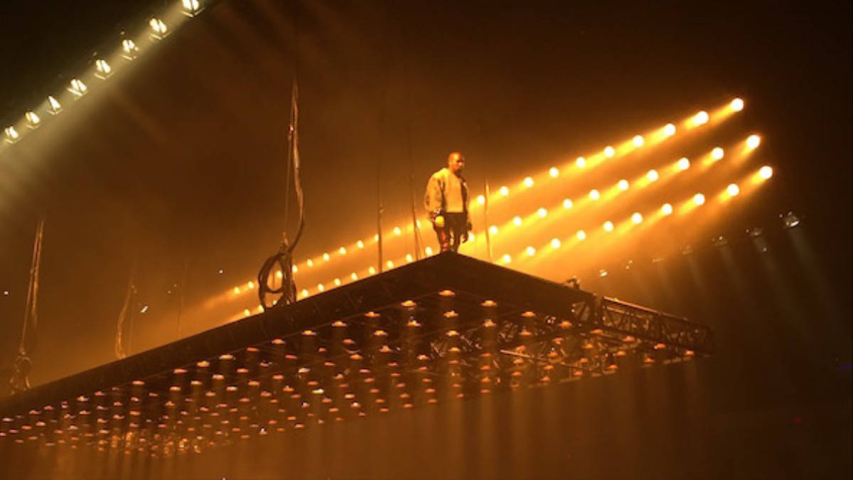 說唱歌手 桌布 壁紙 — 一位孤獨的表演者站在懸掛的舞台上，沐浴在金色聚光燈下，背景是黑暗的音樂會場景。