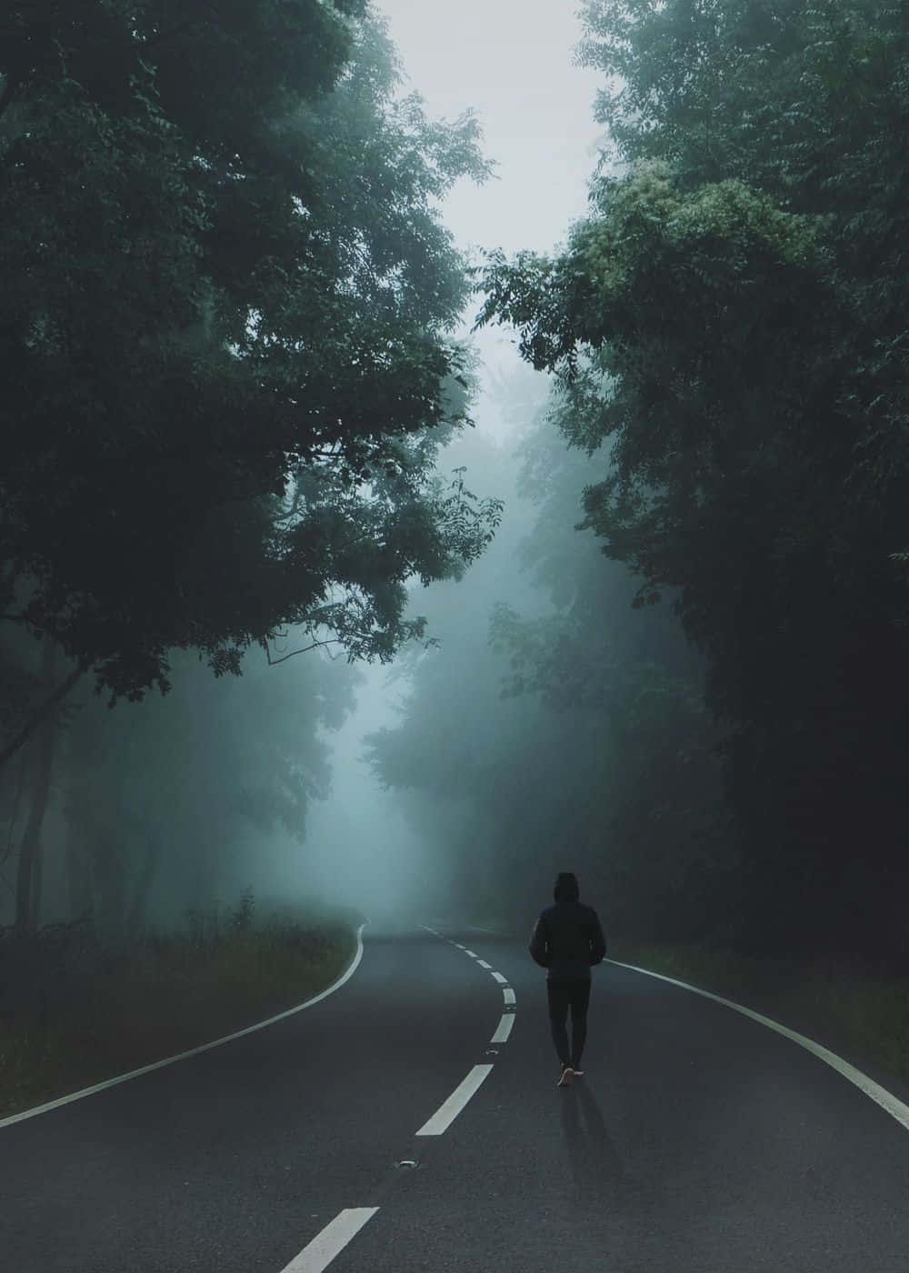 手機 桌布 壁紙 — 一位孤獨的跑者沿著一條霧氣瀰漫的森林道路行走，兩旁是茂密的綠樹，天空朦朧。