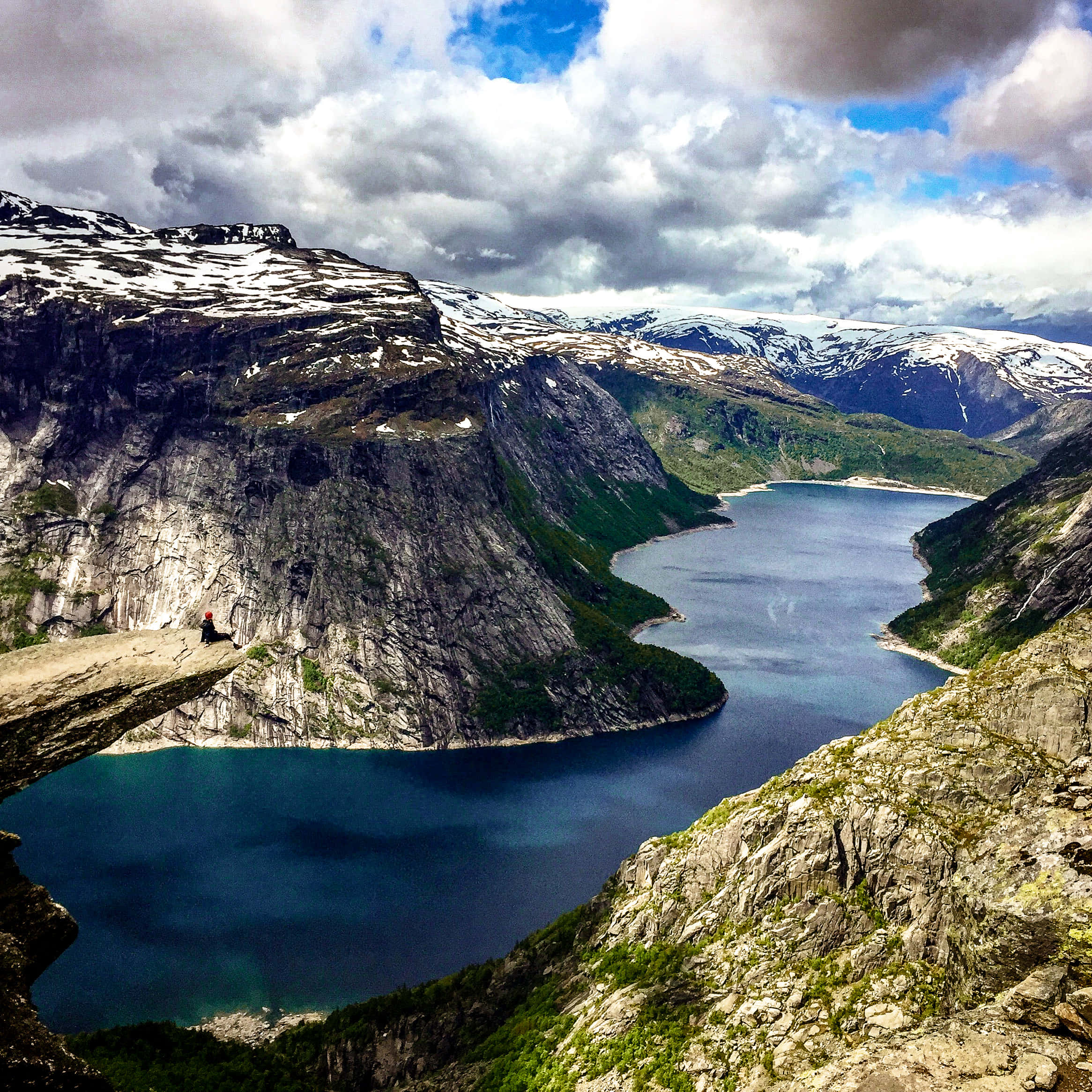 Nature Scenes 桌布 壁紙 — 一位孤獨的遠足者坐在岩石懸崖邊緣，俯瞰著深藍色的峽灣，峽灣位於白雪覆蓋的山脈之間，天空佈滿戲劇性的雲層。