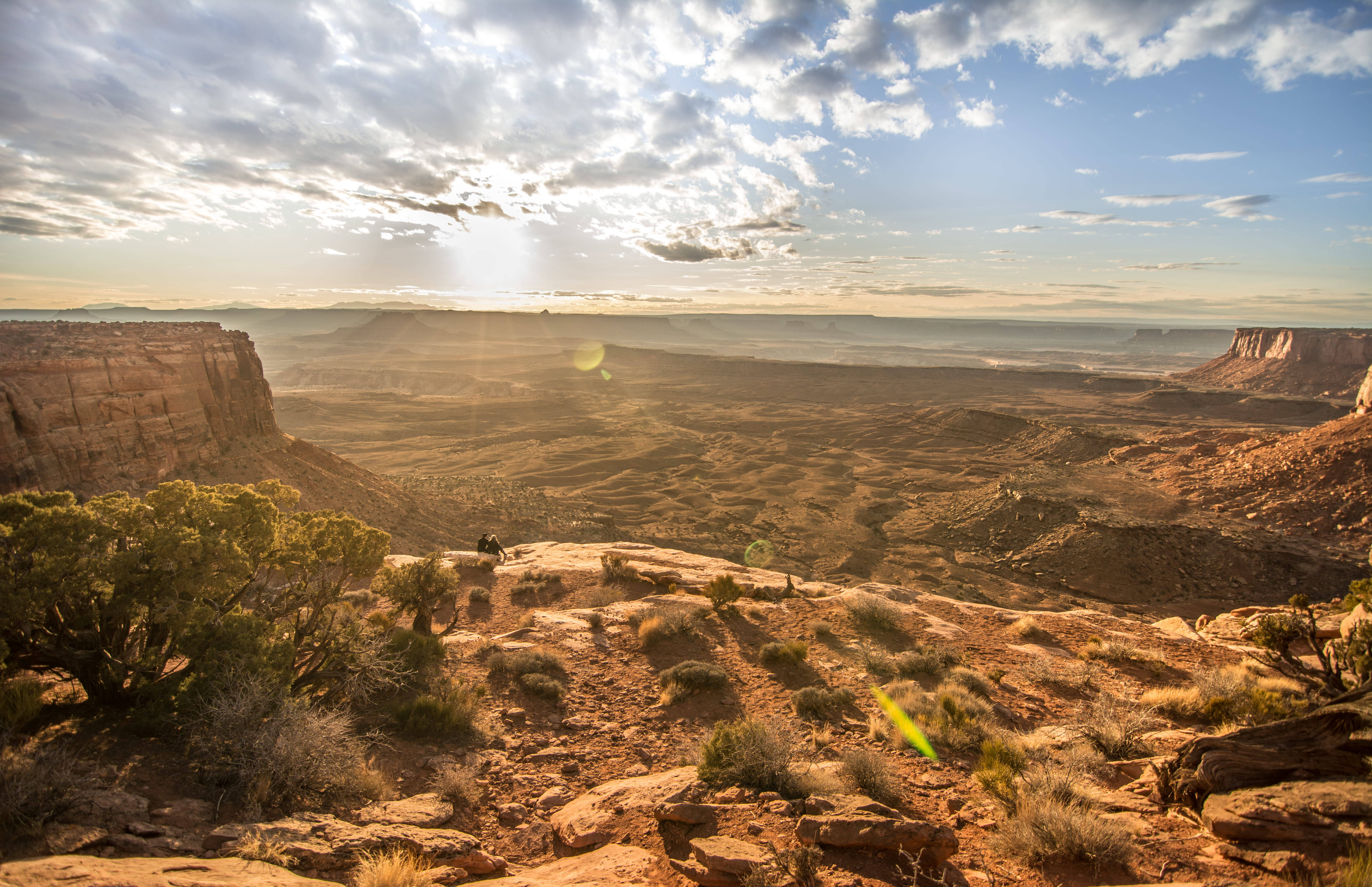 Canyonlands National Park 桌布 壁紙 — 一位孤獨的遠足者坐在陽光普照的峽谷邊緣，俯瞰在戲劇性的多雲天空下廣闊的紅色岩層。