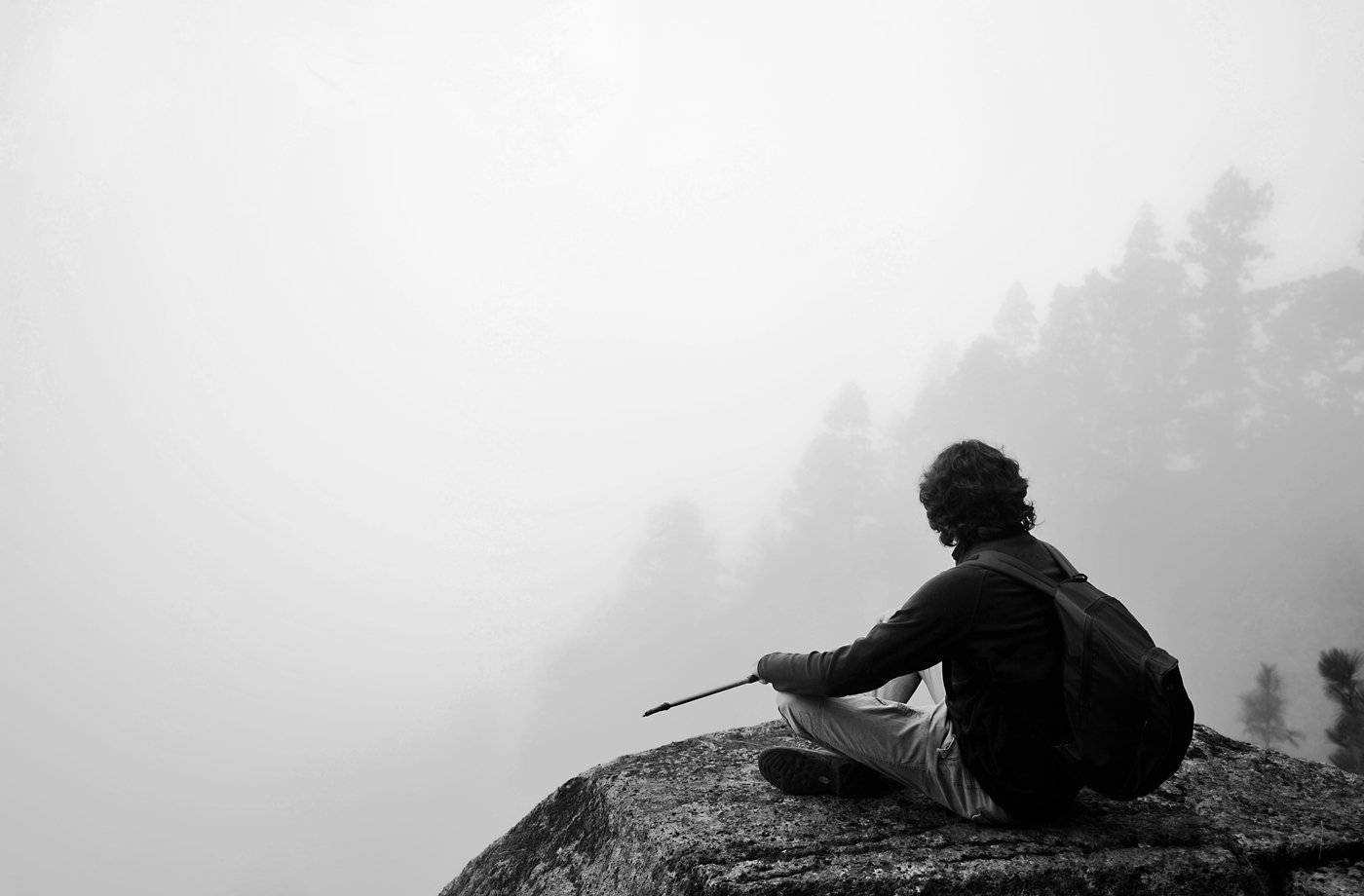 Meditation 桌布 壁紙 — 一位孤獨的遠足者坐在霧氣籠罩的山頂上，手持登山杖，霧氣籠罩著下方的森林。