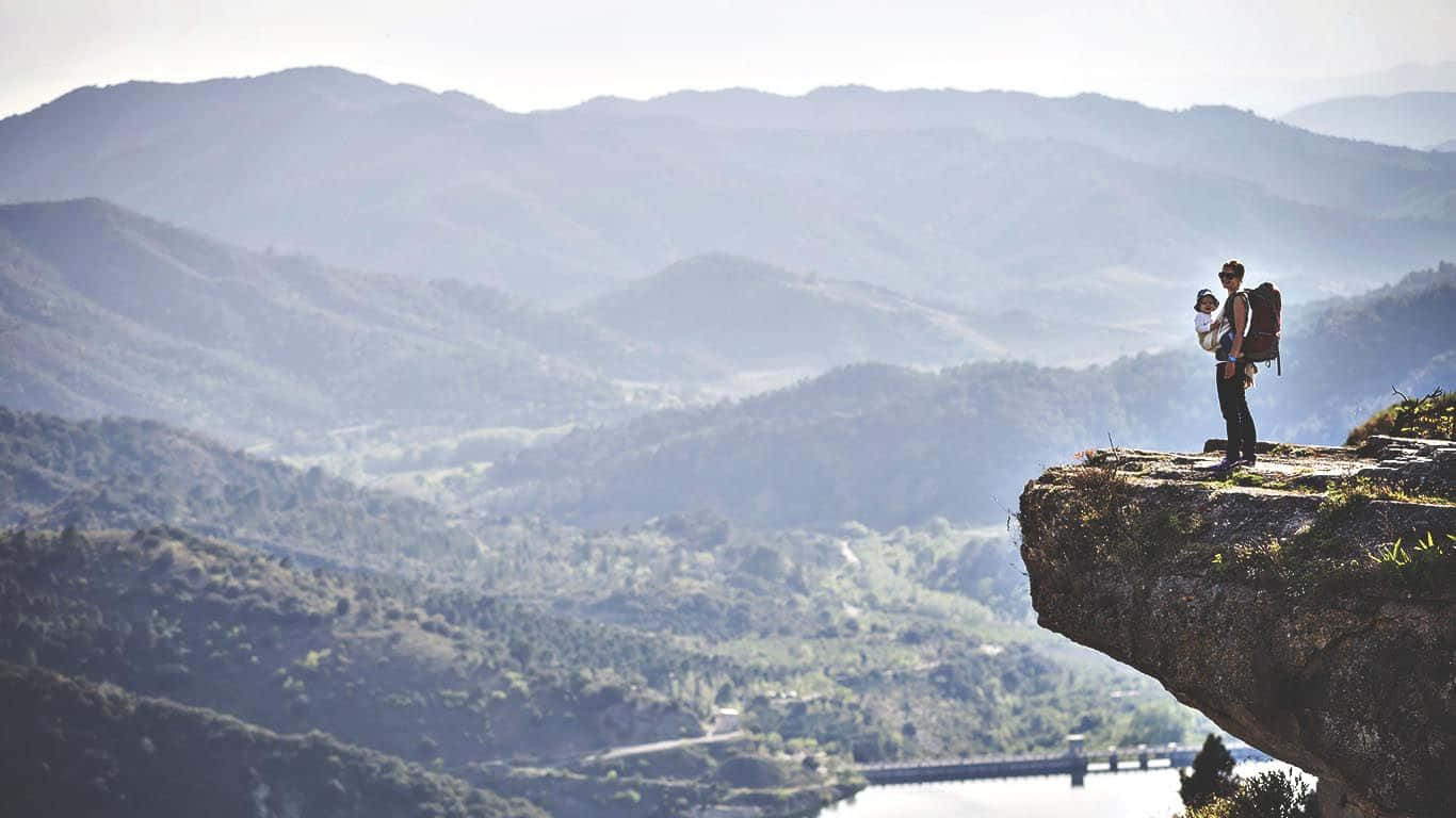 Hiking 桌布 壁紙 — 一位孤獨的遠足者揹著嬰兒在揹架上，站在岩石峭壁頂端，俯瞰著沐浴在柔和晨光中的廣闊山谷。
