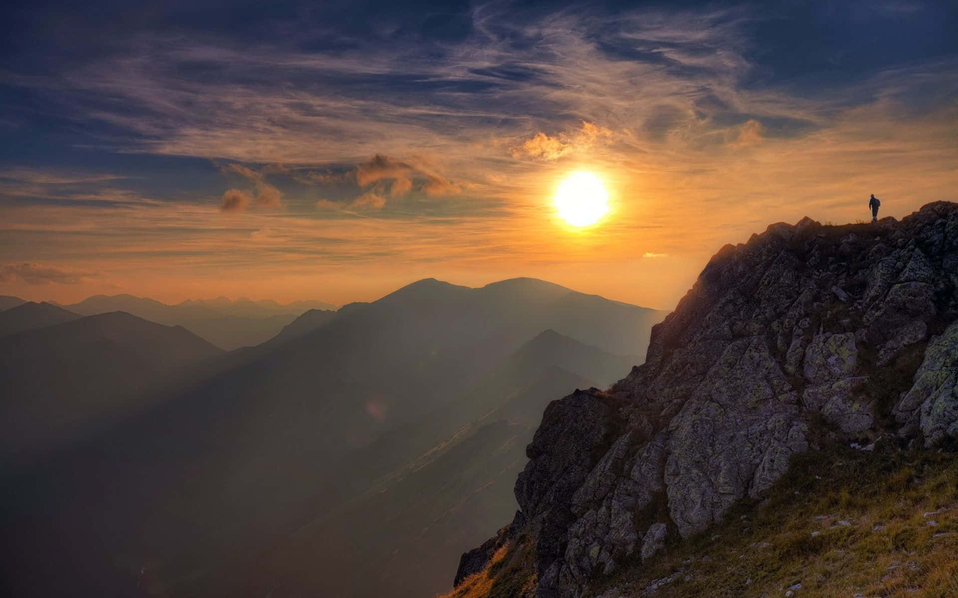 Hiking 桌布 壁紙 — 一位孤獨的遠足者站在崎嶇的山脊上，夕陽在遠峰後落下，將金色光芒灑向山谷。