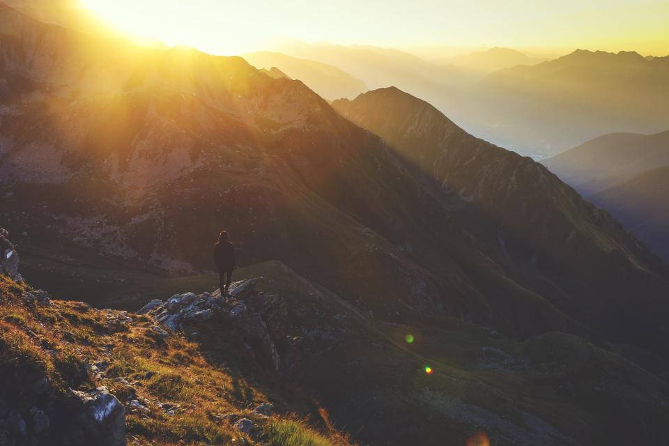 Sad Alone 桌布 壁紙 — 一位孤獨的遠足者站在日出時分陽光普照的山脊上，在發光的山峰和層層疊疊的山谷襯托下形成剪影。