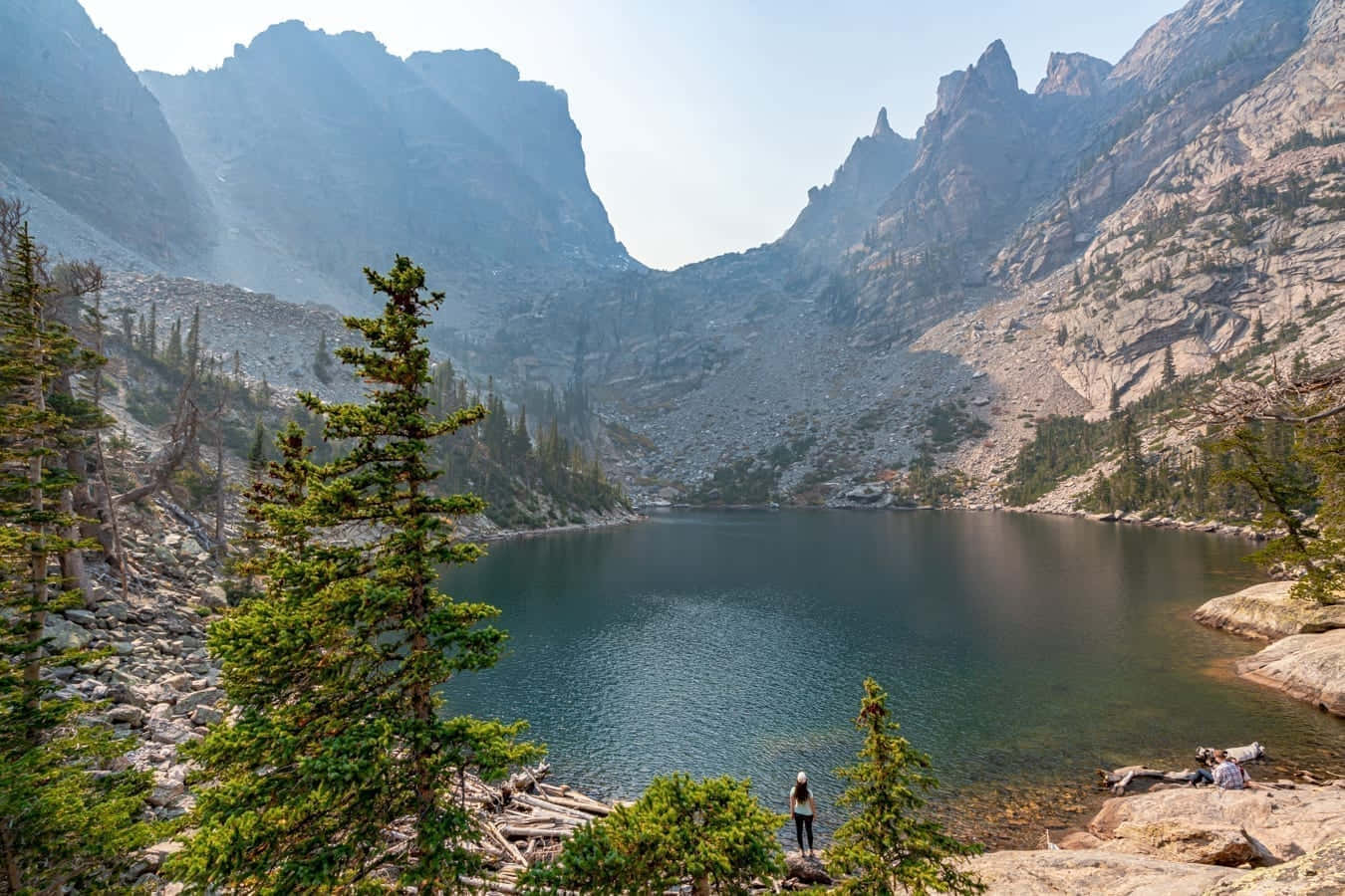 Colorado Mountain 桌布 壁紙 — 一位孤獨的遠足者站在深藍色高山湖的邊緣，周圍是高聳的岩石山峰和常綠樹木，天空朦朧。