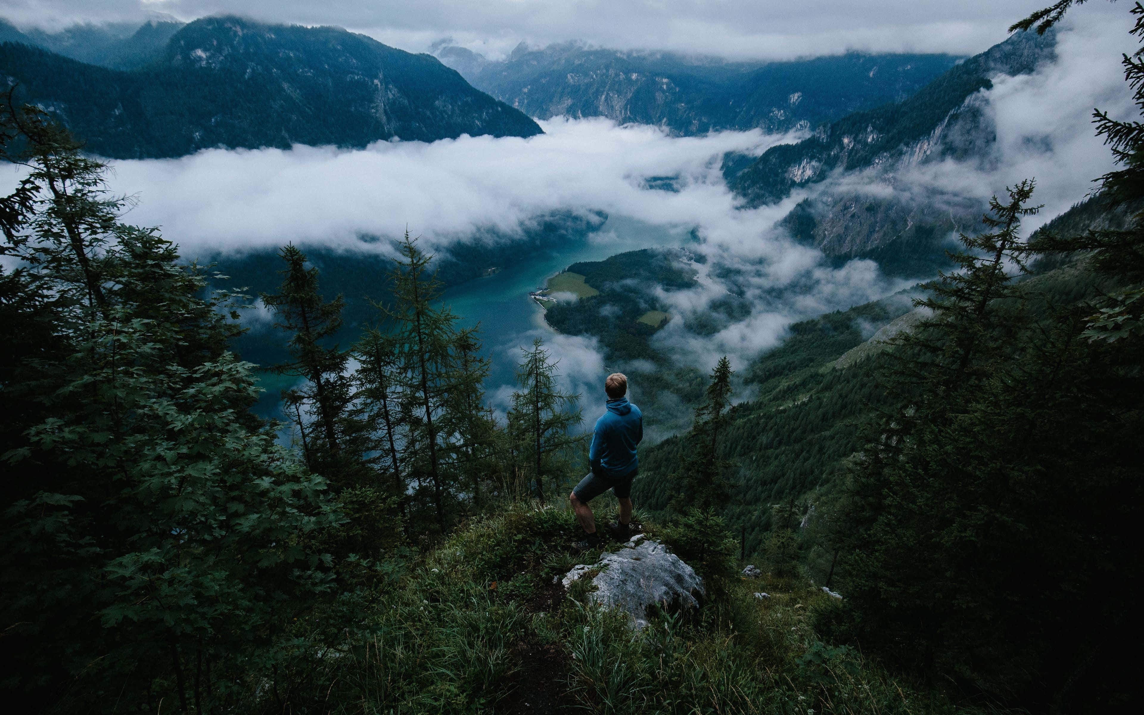 Best Travel 桌布 壁紙 — 一位孤獨的遠足者站在長滿苔蘚的懸崖頂上，俯瞰著一個被高聳的常綠森林和崎嶇山脈環繞的迷霧高山湖泊。