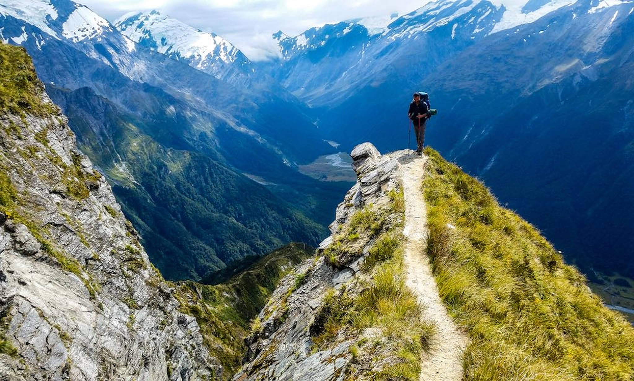登山 桌布 壁紙 — 一位孤獨的遠足者站在長滿草的山脊上，俯瞰著一個深谷，谷中有白雪覆蓋的山峰，天空多雲。