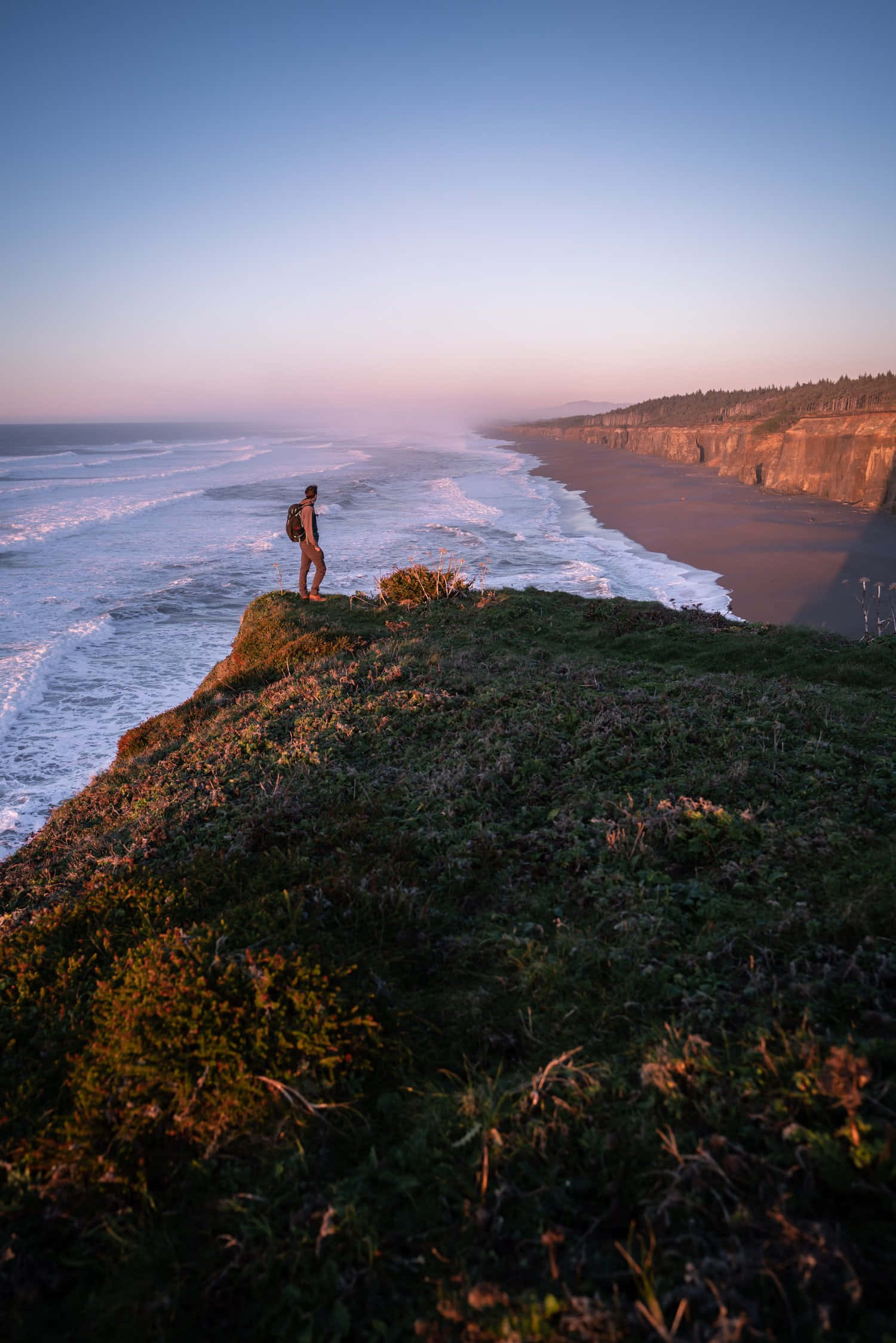 Oregon Coast 桌布 壁紙 — 一位孤獨的遠足者站在長滿草的懸崖邊，俯瞰著日落時分的廣闊海洋，下方波濤洶湧。