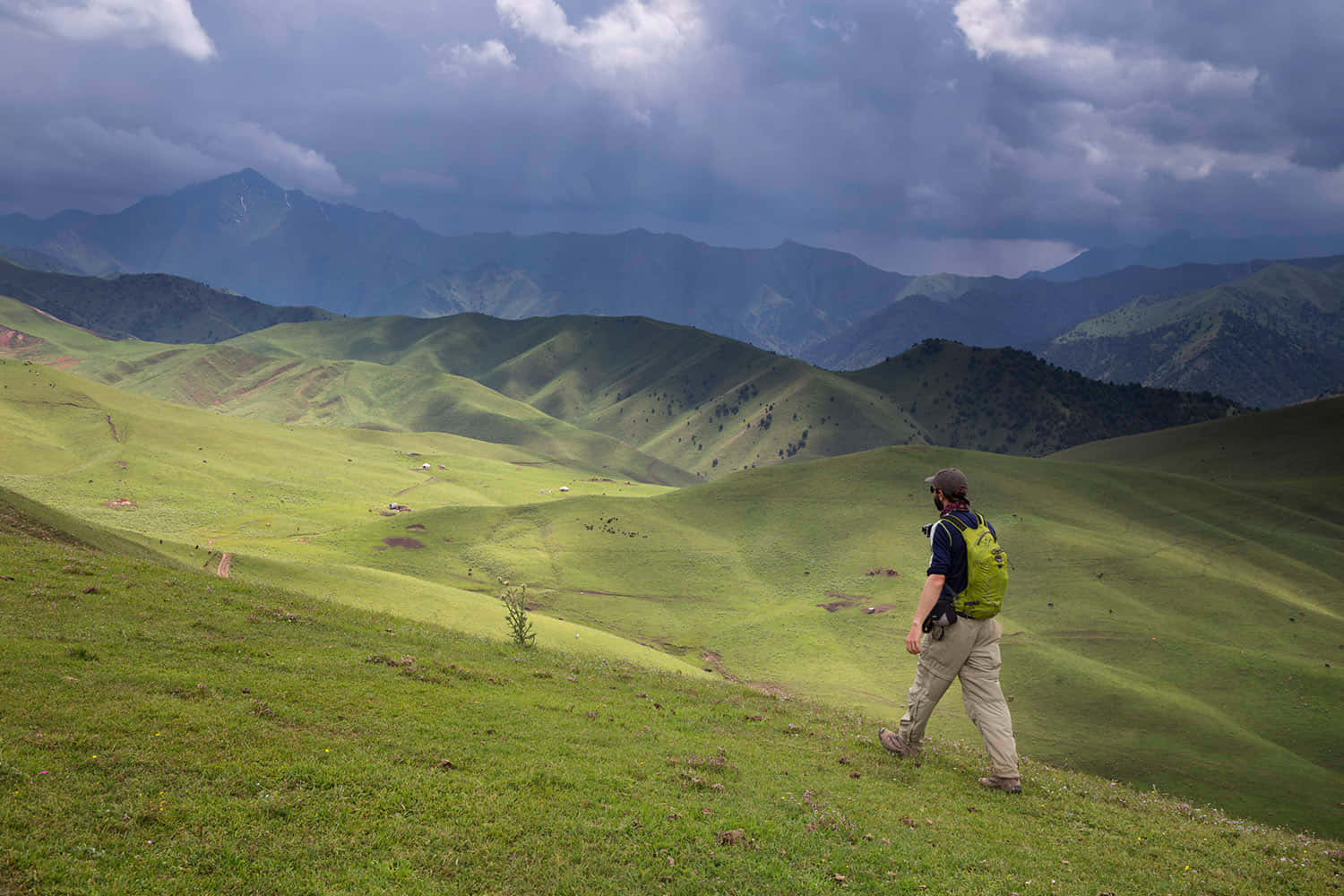 Beautiful Landscape 桌布 壁紙 — 一位孤獨的遠足者背著綠色背包，在戲劇性的多雲天空和遠處山脈下，走過廣闊起伏的綠色草地。